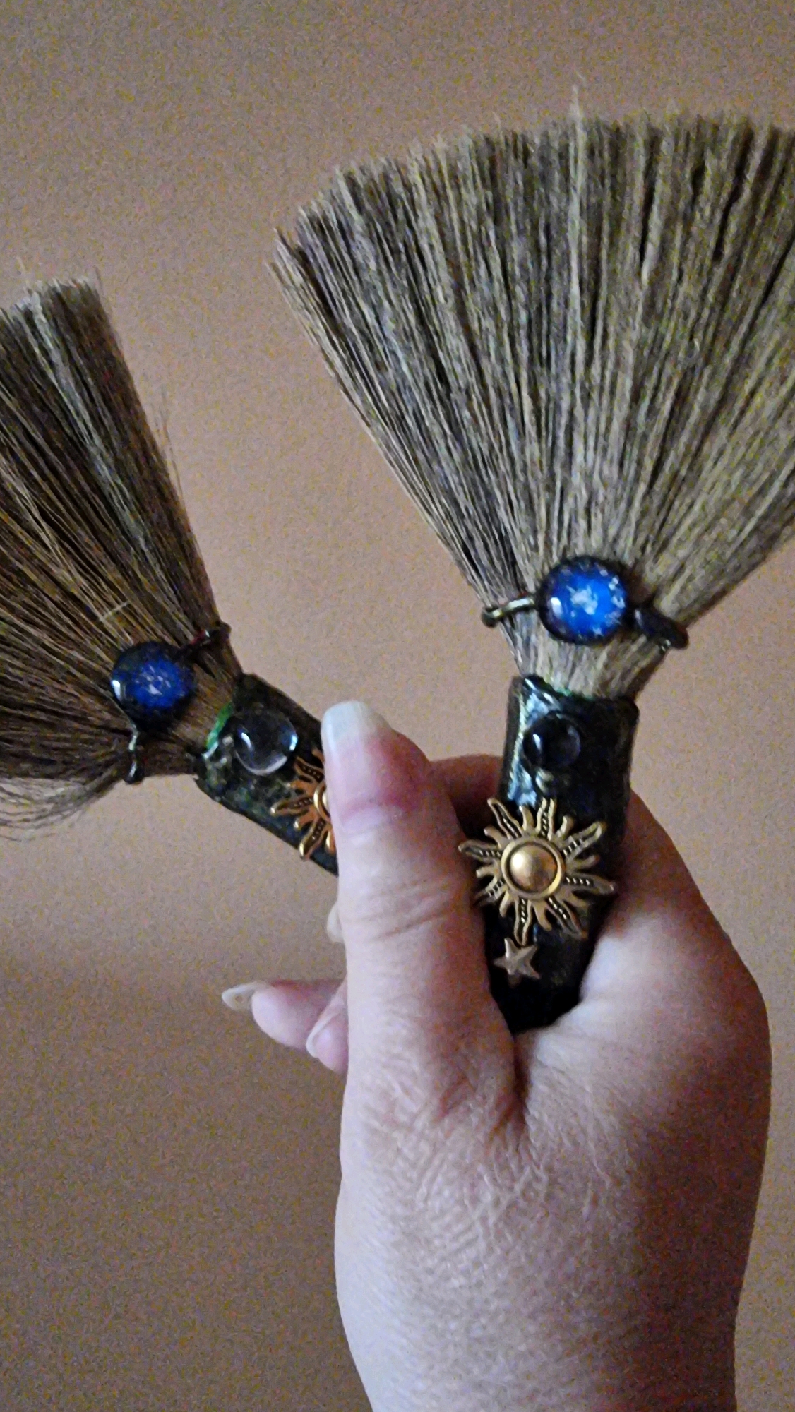 In Sant Joan Les Fonts, Spain, a person's hand is holding two decorative miniature brooms. The brooms have dark handles adorned with celestial motifs, including a blue glass cabochon resembling a starry sky and a sun charm. The bristles of the brooms are tan and feathery. The background is a plain, textured wall in a light brown hue.