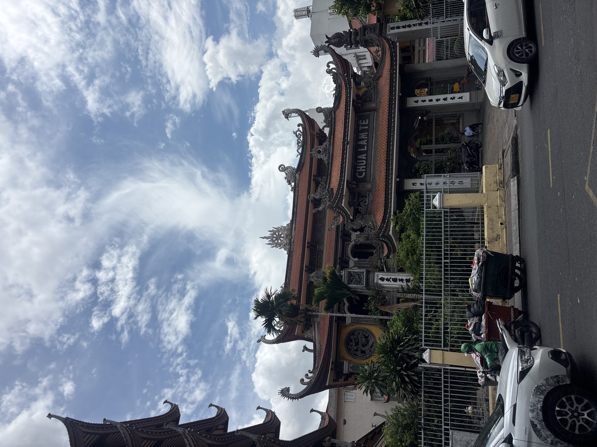 The image shows a street scene in an urban environment, likely in Southeast Asia, given the architecture of the temple and the presence of a motorcycle delivery driver. In the foreground, a white car is parked on the right side of the road, and another white car is partially visible on the left. A man wearing a green uniform and helmet is riding a motorcycle, pulling a cart laden with trash. The cart is being pushed along the sidewalk.
Behind the sidewalk, a traditional temple with ornate, multi-tiered roofs is visible. The temple's facade features intricate carvings and sculptures, including dragons. The name "CHUA LAM TE" is visible above the entrance. A metal fence separates the street from the temple grounds. Lush green foliage and trees surround the temple. The sky is partly cloudy with dramatic, wispy clouds. In the background, modern buildings can be seen peeking out from behind the temple. The overall impression is a blend of traditional and modern urban life.
