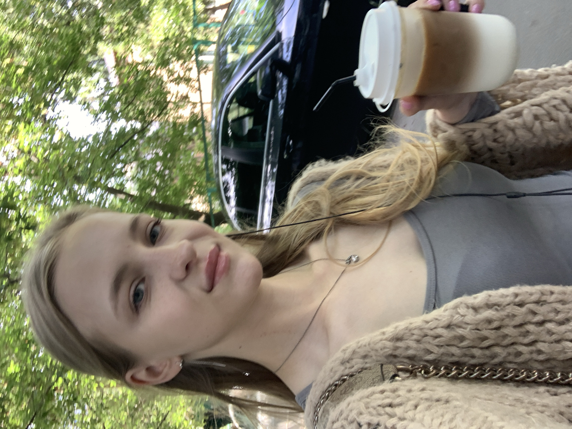 The image is an over-the-shoulder selfie taken outdoors, likely during the daytime as suggested by the natural lighting. The subject is a young woman with blonde hair, wearing a grey top and a beige knit cardigan. She is holding a disposable cup with a white lid and a straw, containing a layered beverage that appears to be coffee with milk or cream. Her facial expression is relaxed and she is looking towards the camera.
In the background, a black car is visible, and lush green foliage suggests an outdoor setting, possibly a street or park with trees. The overall mood is casual and relaxed. The image does not contain any visible text. The mention of "Kyiv, Ukraine" provides a specific geographical context for the scene.