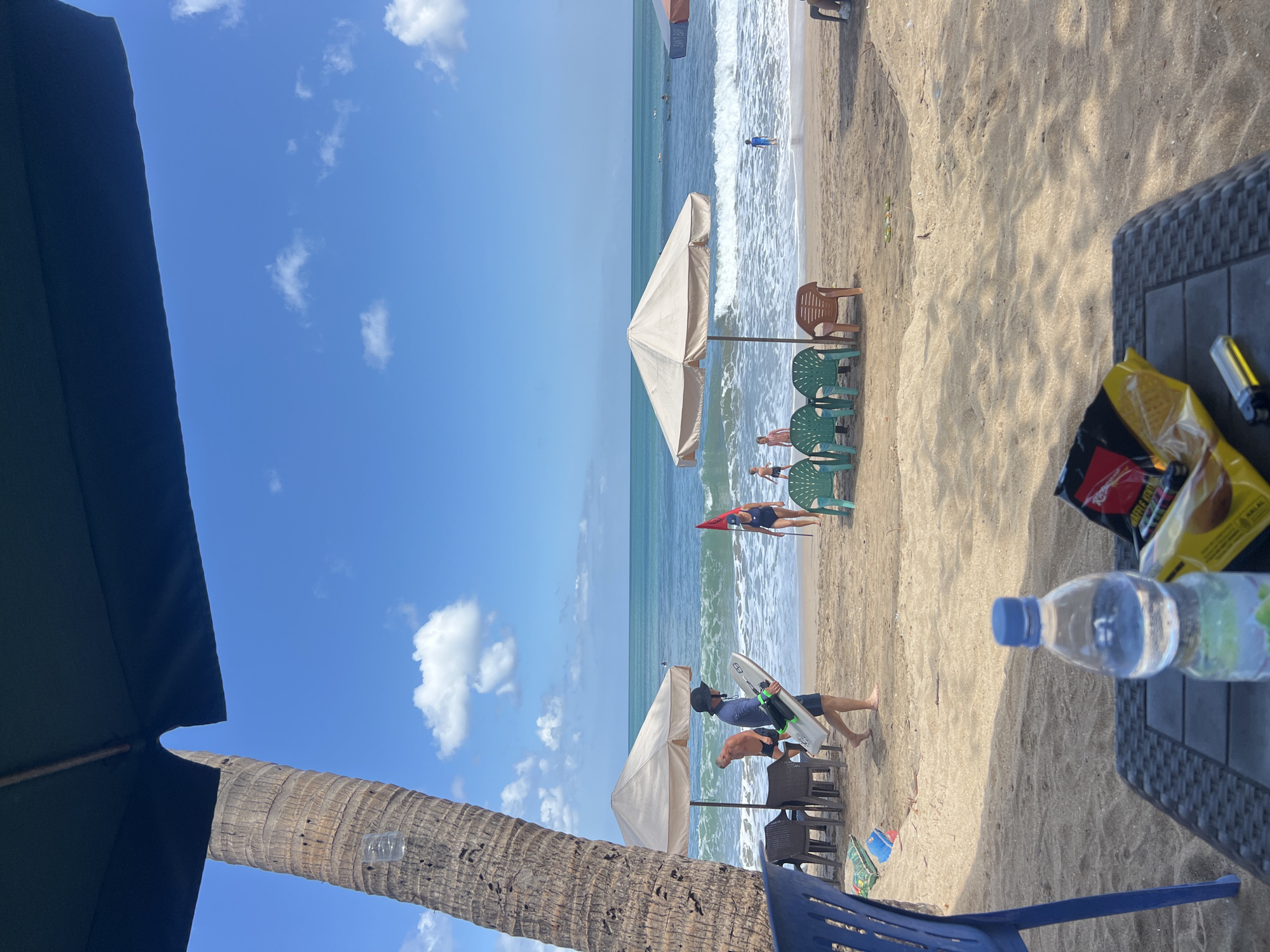 The image captures a sunny day at a beach in Kuta, Indonesia. In the foreground, a table with a bottle of water and a bag of chips is visible. A blue chair is to the left. The beach itself is sandy with some scattered debris. Several beach umbrellas are set up, with chairs beneath them.
Further back, people are enjoying the ocean. One man is walking with a surfboard. Others are in the water or on the sand. The ocean has moderate waves, and several surfers can be seen in the distance. The sky is blue with scattered white clouds. A palm tree trunk is prominent on the left side of the frame, partially obscured by a dark umbrella. A red flag is planted in the sand near the water's edge.