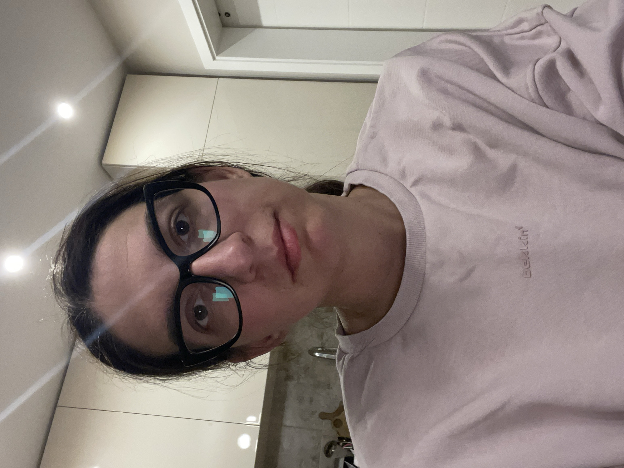 A person is standing indoors, likely in a kitchen, based on the presence of cabinets and a sink with a faucet. The individual is wearing a light pink t-shirt with the word "Bekkin" embroidered on the chest and black-rimmed glasses. The person is looking directly at the camera with a neutral expression. The lighting appears to be from overhead fixtures, casting a bright glow on the upper portion of the image. Without further context, determining the exact location within Plovdiv, Bulgaria, is not possible.