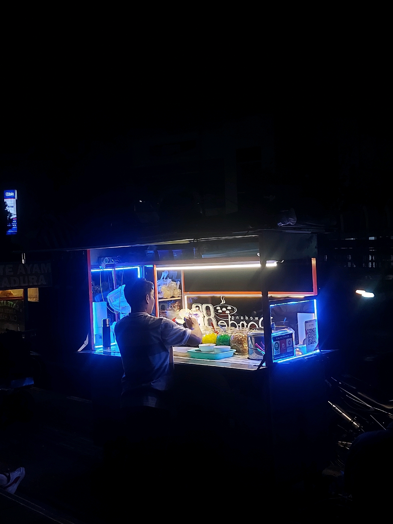 In the city of Wonosobo, Indonesia, at night, a street food stall is illuminated by bright blue and white neon lights. A person, likely the vendor, stands behind the counter, preparing food or drinks. The stall features shelves stocked with various ingredients in jars and containers, including colorful powders and nuts, along with what appears to be food items. A QR code is prominently displayed, suggesting an online ordering option. In the background, there is a sign with text that is partially visible, including "TE AYAM MADURA". The overall scene depicts a typical nighttime scene at a bustling Indonesian street food vendor.