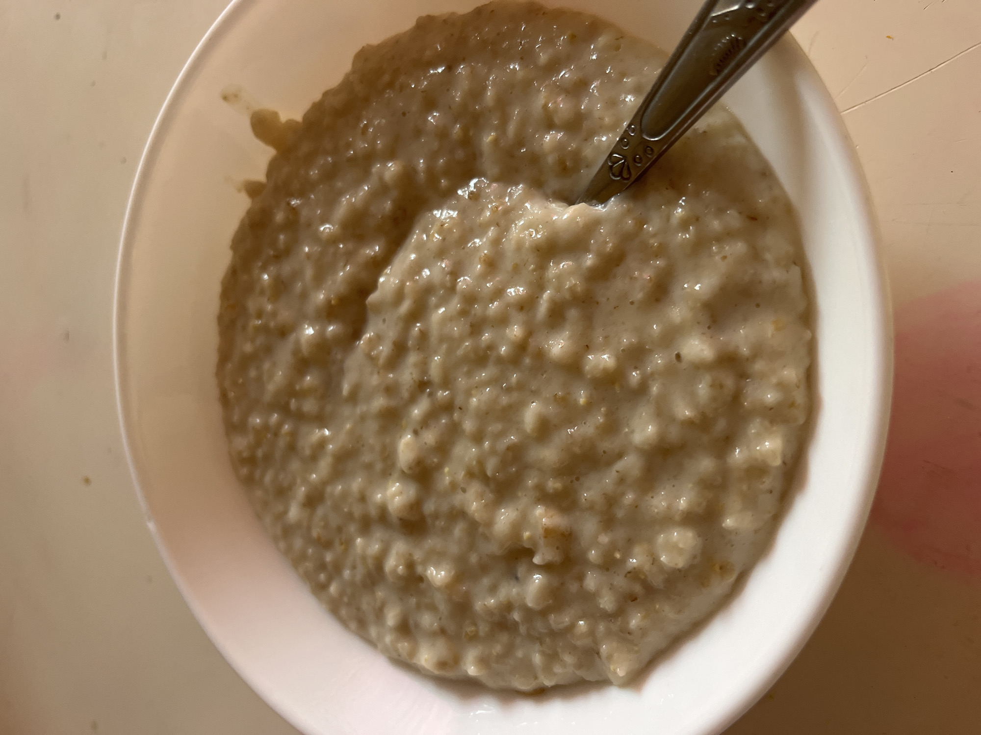 The image shows a close-up, overhead view of a white bowl filled with oatmeal. A spoon with a decorative pattern on its handle is partially submerged in the oatmeal on the right side of the bowl. The oatmeal appears creamy and has a slightly textured surface. The bowl is set on a light-colored, possibly plastic surface. There is no visible indication of the location being Tarnivtsi, Ukraine, or any specific city.