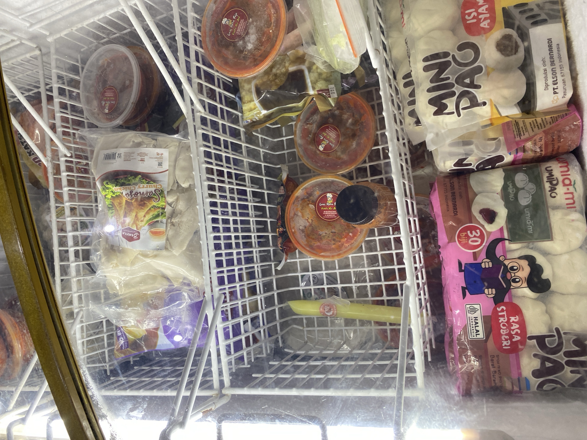 The image depicts the interior of a commercial freezer filled with various frozen food products. The freezer shelves are made of white wire grating. On the top shelf, there are several clear plastic containers with reddish-brown contents, possibly sauces or dips. To the left of these, a package of what appears to be dim sum or dumplings is visible, with a label featuring a cartoon image of a chef and text in Indonesian.
The middle shelf contains more clear plastic containers with similar reddish-brown contents. A bright yellow cylindrical item, possibly a frozen treat or a food item, stands upright in the center of this shelf. Behind it, more packaged food items are visible.
The bottom shelf is crowded with several large, transparent plastic bags of frozen food. One prominent bag is pink and features a cartoon character, with text in Indonesian such as "Rasa Stroberi" (Strawberry Flavor) and "30 biji" (30 pieces). Another bag, labeled "MINI PAO," contains small steamed buns and has text indicating "ISI AYAM" (Chicken Filling). Other bags contain food items with Indonesian text.
The overall environment appears to be a retail setting, likely a supermarket or a specialized food store in Payatieng, Indonesia, given the presence of Indonesian text on product packaging. The lighting suggests the photograph was taken inside the freezer, with some light reflecting off the clear plastic packaging and the freezer's glass door. There are no people or animals visible in the image. The items are neatly arranged on the shelves, indicating a well-stocked and organized freezer.