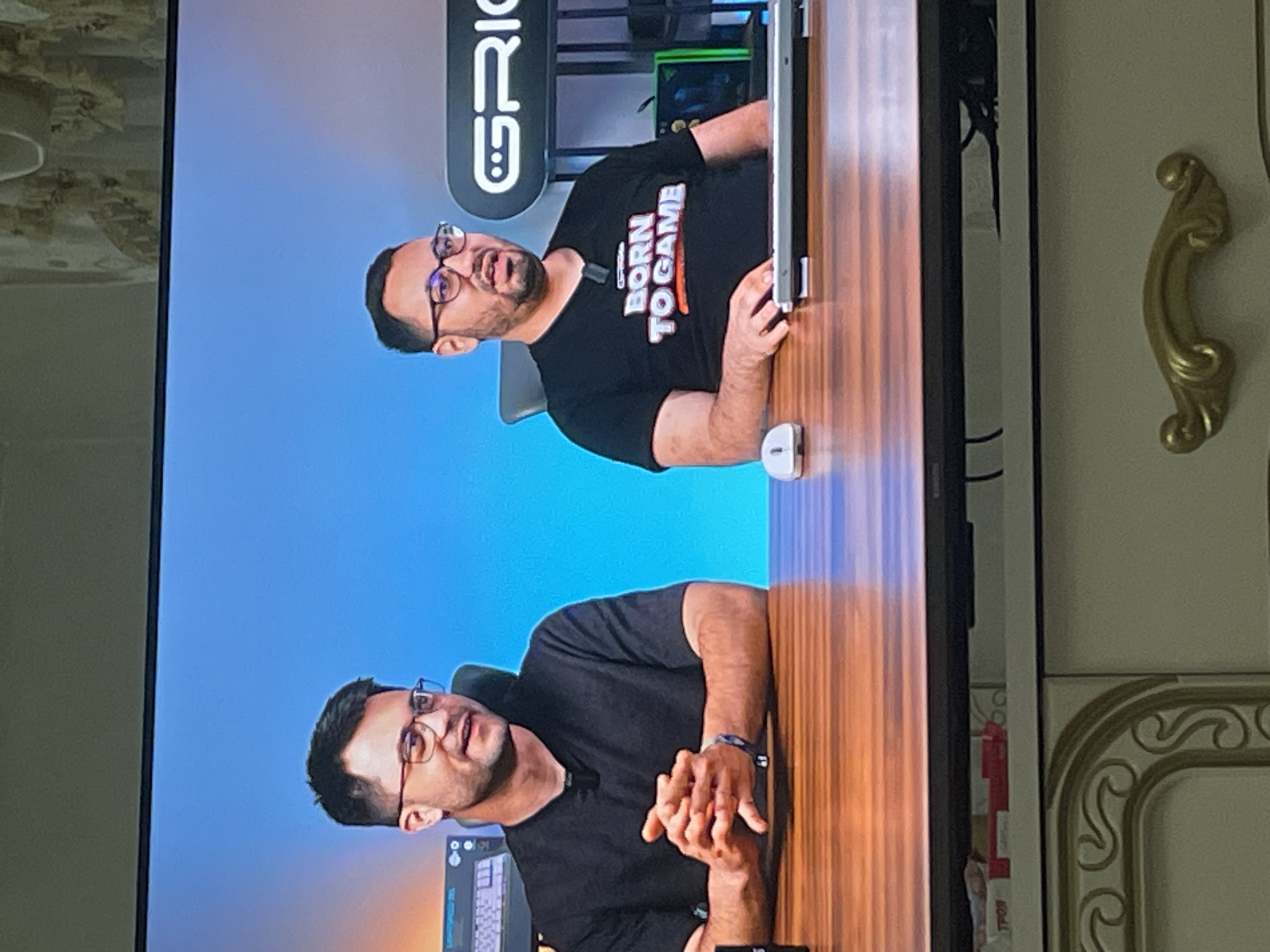 A television screen, originally rotated 90 degrees, displays two men seated at a wooden desk against a light blue background.
The man on the left (when the image is correctly oriented) wears a black short-sleeved shirt and glasses. His hands are clasped on the table, and he has a dark watch on his left wrist. A microphone is clipped to his shirt.
The man on the right wears a black short-sleeved t-shirt with the text "BORN TO GAME" in white and orange/red letters. He also wears glasses and has a microphone clipped to his shirt. His hands rest on an open laptop or portable screen. A white computer mouse is visible near his right hand.
In the background of the TV screen, behind the men, there are shelves with what appears to be gaming equipment, including a green PC case. A logo with stylized letters, possibly "GRIG" or "G RIG," is visible on the wall. The men appear to be engaged in a discussion or presentation, suggesting a video podcast, stream, or interview.
Outside the television frame, to the right, a decorative gold-colored handle is attached to light-colored furniture, possibly a cabinet or door, featuring ornate carvings or molding. To the left of the TV, a textured, light-colored wall is visible. Some cables and a white structure are partially seen behind the TV. A red and white object, possibly a package, is also partially visible behind the right side of the screen.
The image captures an indoor setting in Ihtiman, Bulgaria, where someone is watching content on a TV screen.