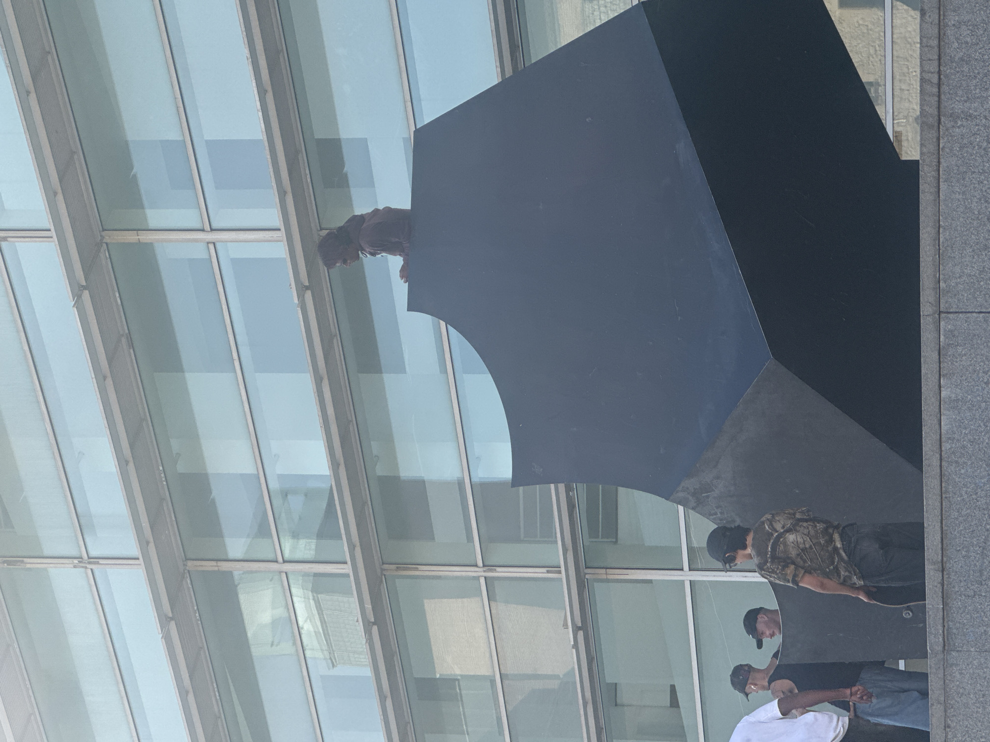 The image, rotated 90 degrees clockwise, captures a dynamic urban scene centered around a large, dark, geometric sculpture and a group of young men. Shot from a low angle, the composition highlights the interplay between the sculpture, the people, and a modern glass building.
At the upper left, a person wearing a reddish-brown top and dark, curly hair is positioned on the top edge of the large, abstract, dark gray sculpture, seemingly looking down or in the process of climbing. This sculpture features sharp angles and a distinctive concave curve on one of its faces.
To the lower right, three other young men stand on a gray, paved surface next to the sculpture. The most prominent figure, facing left, wears a black baseball cap, a camouflage-patterned t-shirt, and dark pants, holding a skateboard under his arm. Behind him, another individual in a black cap and dark t-shirt looks towards the sculpture. A third person, partially visible, wears a white t-shirt and blue jeans. Their collective posture suggests a casual gathering.
The background features a striking modern building with a facade entirely made of large, reflective glass panels, creating strong diagonal lines and reflecting the bright blue sky and distant structures. The clear reflections indicate bright daylight. The overall atmosphere is relaxed, set in what appears to be a contemporary public space. There is no visible text in the image. The architectural style is consistent with an urban environment in a city like Barcelona.