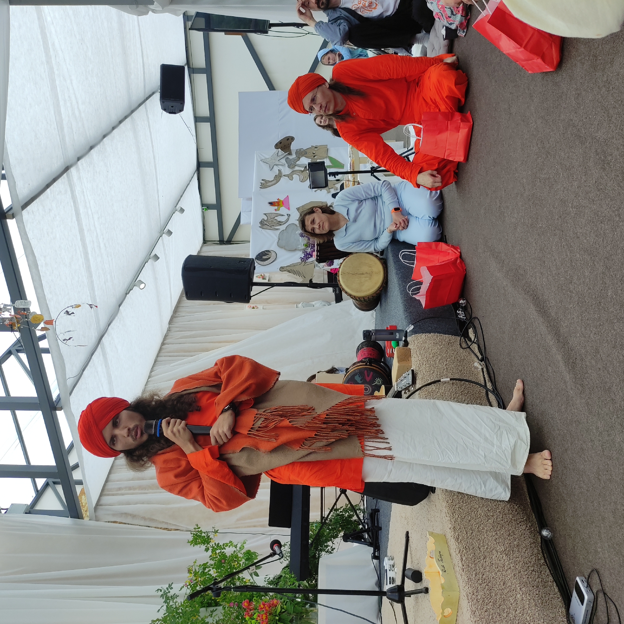 In Zhirnosovo, Russia, a man wearing an orange turban and robe stands on a raised platform, speaking into a microphone. He has long, dark curly hair and a light brown scarf draped around his shoulders. To his left, a keyboard and microphone stand are visible. To his right, two people are seated on the floor, facing away from the viewer. The person on the left, a woman in a light blue tracksuit, looks towards the speaker. The person on the right, dressed in an orange robe and turban, looks to the side. Several red gift bags are scattered on the ground. Behind the seated individuals, a large white cloth forms a canopy or backdrop. Various drums and musical instruments are also present on the stage. The setting appears to be an outdoor event, possibly a cultural gathering or festival.