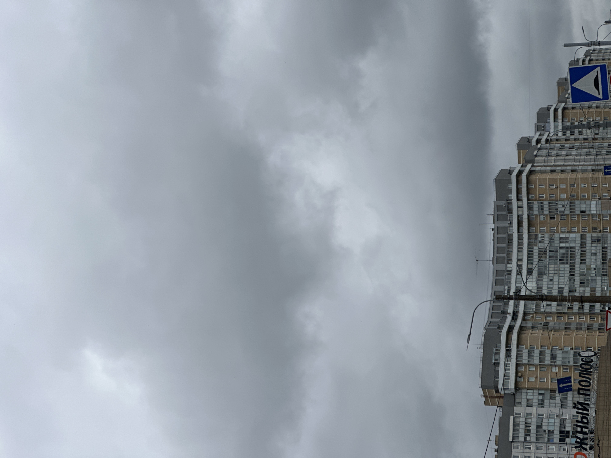 The image shows a cloudy, overcast sky with a portion of a modern residential building. The building is tall and multi-story, with a facade featuring a mix of beige and gray panels, numerous windows, and balconies. A street lamp is visible attached to a pole in front of the building. Two blue and white road signs are also present, one with a symbol that appears to be a warning for a speed bump or uneven road surface, and another with an arrow pointing to the left. Below the signs, a portion of a large sign with white Cyrillic text is visible on the building itself, with the words "ЖНЫЙ ПОЛЮС" clearly legible. The overall atmosphere is somber, likely due to the heavy cloud cover, suggesting it is daytime but with poor visibility. The context provided indicates the location is Saint Petersburg, Russia. There are no people visible in the frame.