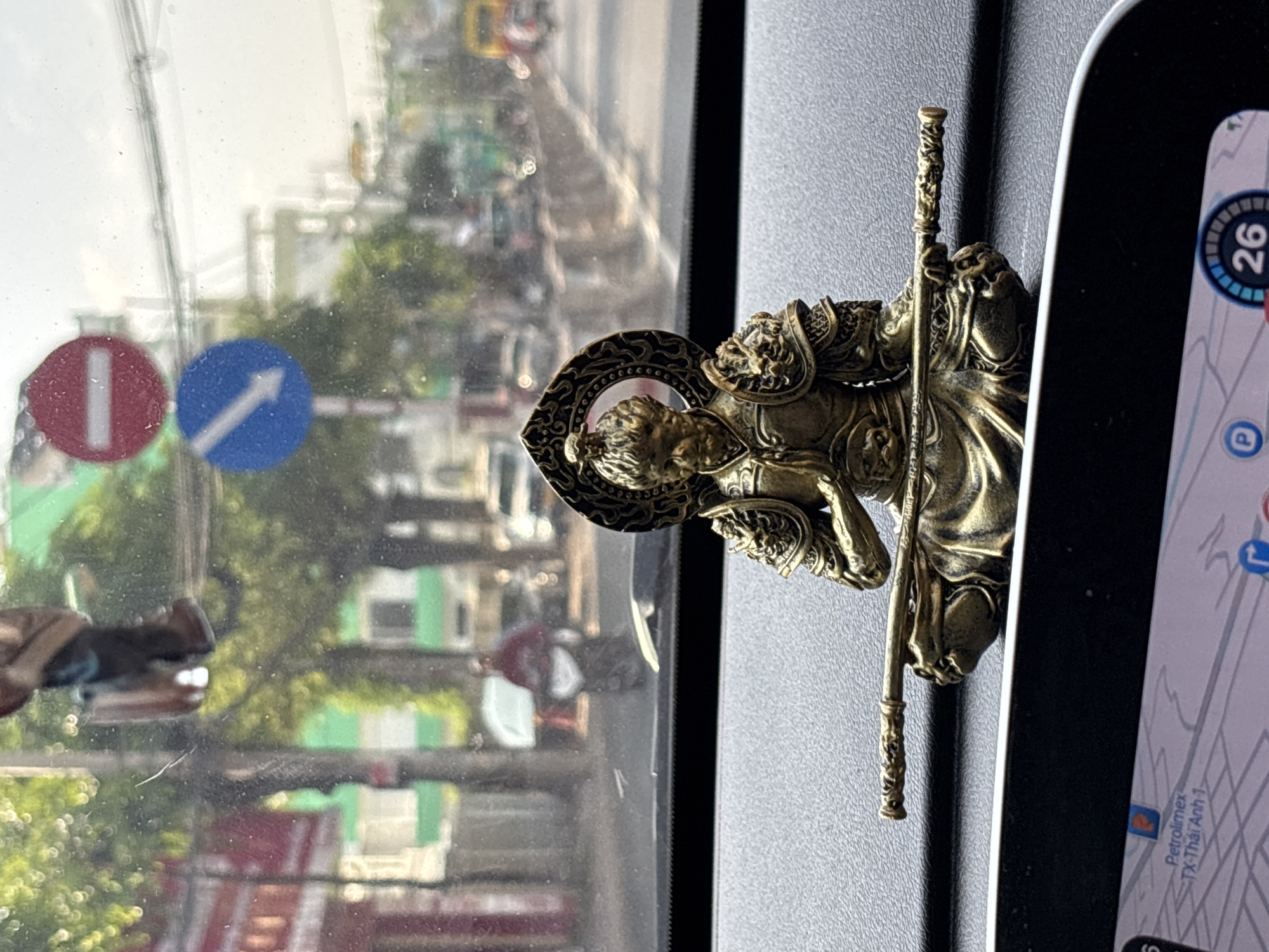 In Nha Trang, Vietnam, a bronze figurine of Sun Wukong, the Monkey King from Chinese mythology, is placed on the dashboard of a car. The figurine is seated with a golden staff held horizontally across its lap. The background visible through the car's windshield shows a street scene with trees, buildings, traffic, and road signs. One sign is a red circle with a white horizontal bar, indicating "no entry" or "do not enter." Below it is a blue circular sign with a white arrow pointing down and to the left, signifying "turn left." The dashboard also features a GPS navigation screen at the bottom of the image, displaying a map with location information.