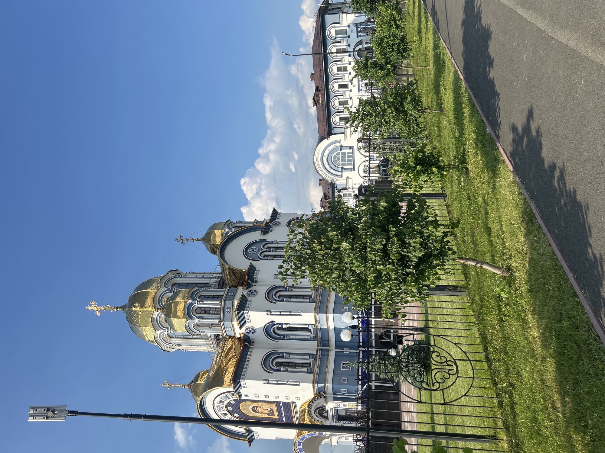 The image shows a white and blue Orthodox church with golden domes, located in Kemerovo, Russia. The church has arched windows and ornate architectural details. In front of the church, there is a black metal fence, a well-maintained lawn, and several small trees. A paved path runs along the edge of the lawn. A tall street lamp with a fixture on top stands to the left of the church. In the background, another building with a brown roof and white and blue facade is visible. The sky is blue with scattered white clouds. There are no people or animals visible in the image.
