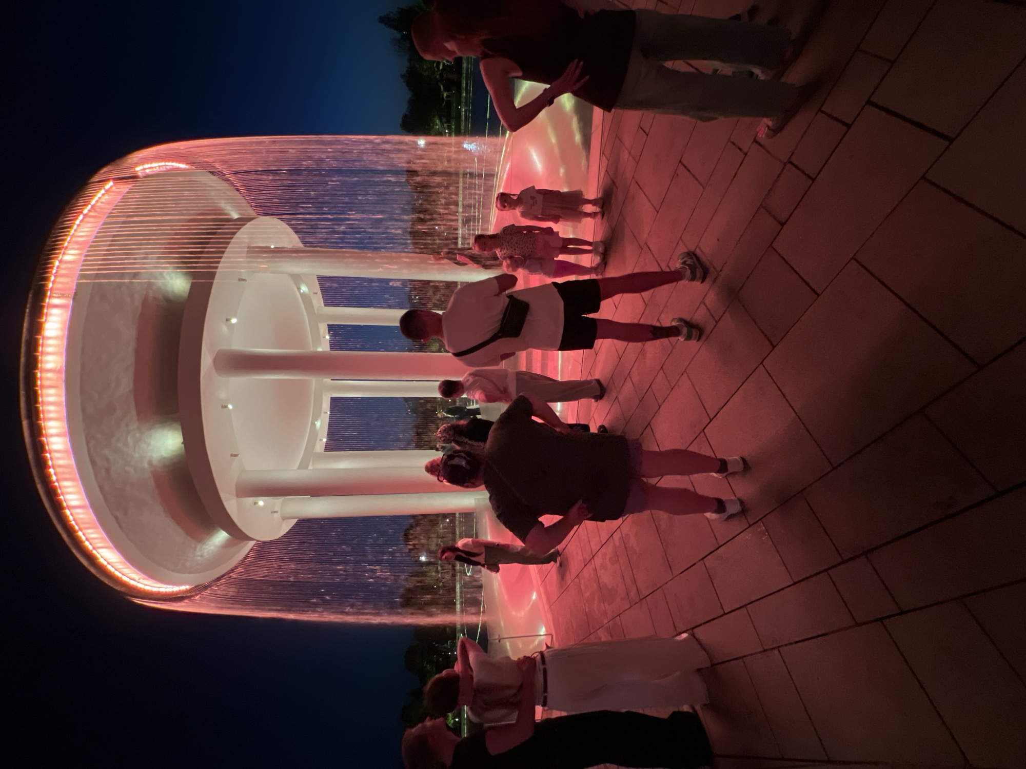 The image shows a group of people standing near a large, illuminated fountain at night. The fountain features a circular structure with vertical water streams that are lit with pink and red lights, creating a vibrant and dynamic visual effect. Several individuals, including adults and children, are visible in the foreground and background, appearing to observe or interact with the fountain. The ground is paved with square tiles, and the overall setting suggests an outdoor public space, possibly a park or plaza. The time of day is clearly night, indicated by the dark sky and artificial lighting. The atmosphere seems relaxed and enjoyable, with people gathered to experience the illuminated water display. While the exact location isn't explicitly stated in the image, the provided context places it in Krasnodar, Russia. No visible text is present in the image.