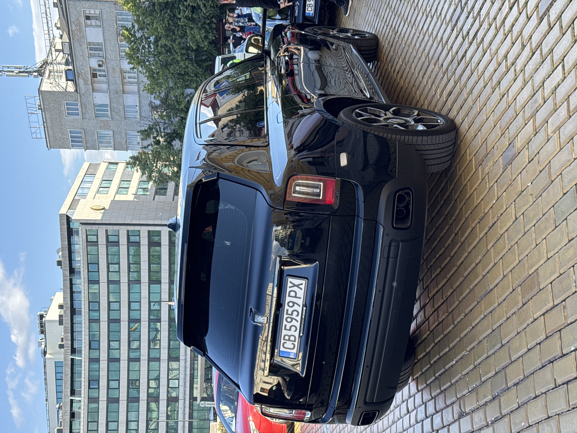 A black Rolls Royce Cullinan is parked on a brick-paved street in Sofia, Bulgaria. The license plate reads "CB 5959 PX". Modern office buildings can be seen in the background. Several people are standing in the distance.