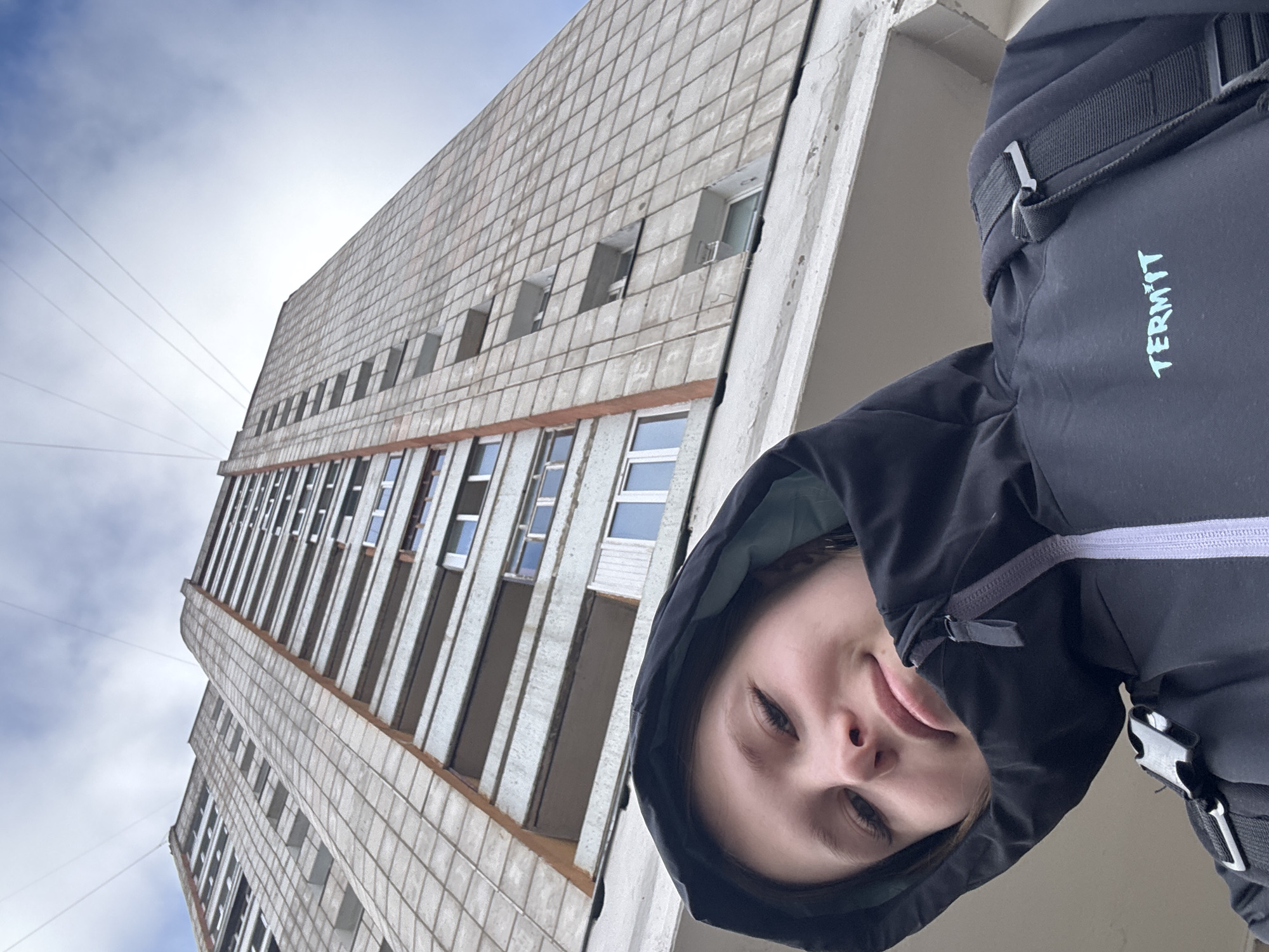 A low-angle shot shows a building and a person in Chaikovsky, Russia. The building is tall and gray, with a grid-like facade and rectangular windows arranged vertically. Power lines stretch across the sky in the upper-left corner. The person in the lower-right is wearing a dark hooded jacket with the word "TERMIT" printed vertically in turquoise. The face is visible, showing fair skin and light eyes.
