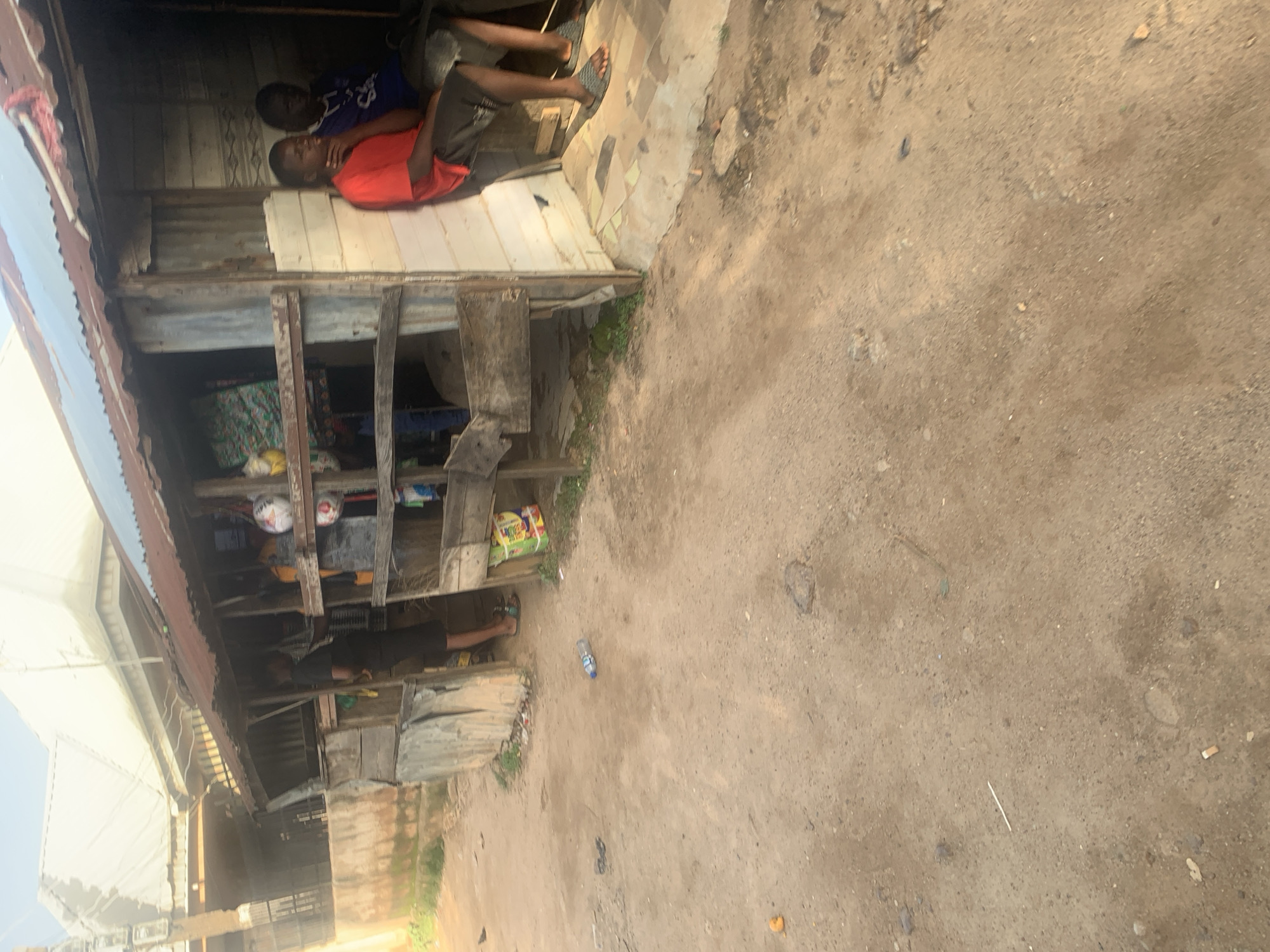 In a dusty, open area in Jos, Nigeria, a makeshift wooden stall with a corrugated metal roof stands. Two young males are seated in the open stall. The male on the left wears a red t-shirt and dark shorts, with his hand on his chin. The male on the right wears a blue t-shirt and dark shorts. A person wearing a black t-shirt and black shorts stands inside the stall, facing away from the camera, with their back to a shelf that displays various items, including what appears to be a soccer ball and bags of goods. In front of the stall, a discarded plastic water bottle lies on the dirt ground. The scene depicts a humble marketplace or roadside stall environment.