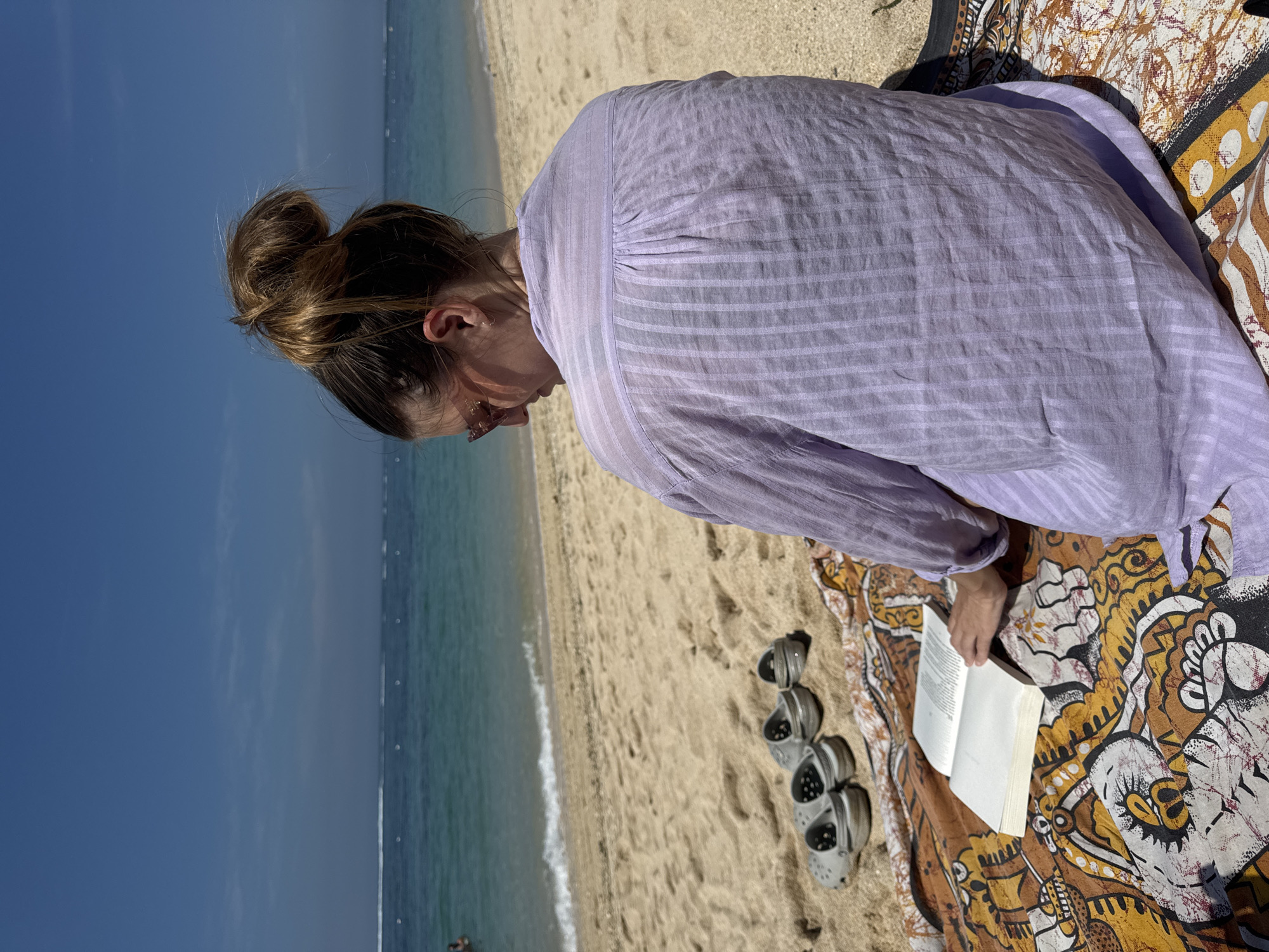A woman sits on a patterned cloth on a sandy beach, reading a book. She wears a long-sleeved, light purple striped shirt and has her hair pulled up in a bun. A pair of gray clogs rests near her on the sand. The ocean and a clear blue sky are visible in the background. The location is in the city of Unknown, Unknown.