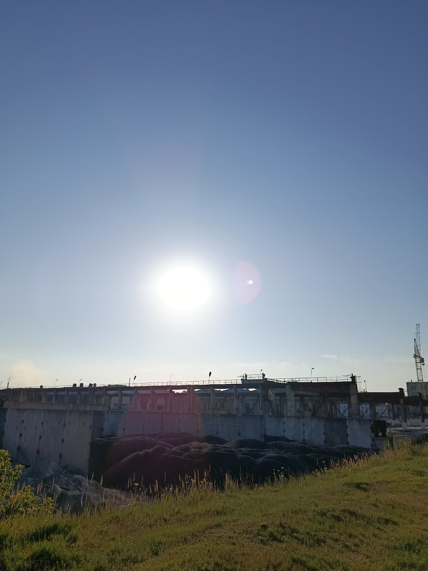 The image depicts a view of the Svietlahorsk Hydroelectric Power Station in Svietlahorsk, Belarus. The foreground consists of a grassy embankment. In the midground, a large concrete structure, which appears to be part of the power station, is visible. Piles of dark material, possibly coal or gravel, are situated in front of the concrete structure. Above the structure, the sky is clear and blue, with the sun prominently featured, creating lens flare. In the background, a construction crane is visible on the right side of the frame, suggesting ongoing work or development. There is no clear indication of people or specific actions taking place. The overall scene captures an industrial landscape under a bright, sunny sky.