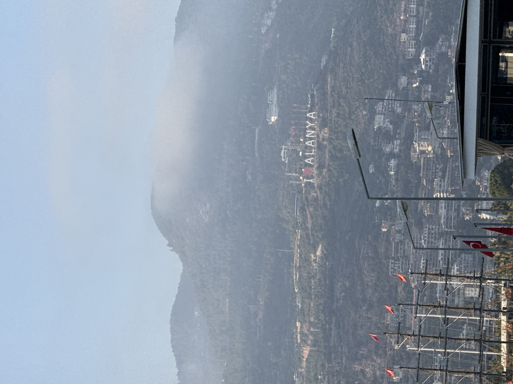 This is a vertical, medium-shot photograph taken from an elevated vantage point overlooking a coastal city and its mountainous surroundings. The scene is bathed in a hazy, possibly early morning or late afternoon light, contributing to a soft, diffused atmosphere. The weather appears to be clear but with some atmospheric haze or mist, particularly obscuring the distant mountains.
The dominant subject is the city of Alanya, Turkey, nestled at the foot of a large, tree-covered mountain. On the mountainside, a large sign spells out "I ALANYA" in white, blocky letters. Several red flags with white crescents and stars, presumably Turkish flags, are visible, some flying from poles near the sign and others adorning what appear to be moored sailboats in the foreground. These sailboats have multiple masts and rigging, suggesting a harbor or marina.
The city itself is characterized by a dense collection of white and light-colored buildings, densely packed together along the slopes and at the base of the mountain. These buildings appear to be hotels or residential structures, typical of a Mediterranean resort town. In the distance, the mountains fade into a soft, atmospheric haze, with the outlines of structures only faintly discernible.
In the immediate foreground, parts of a modern building or structure are visible on the right side of the frame, with dark window frames and a lighter-colored façade. Streetlights and lamp posts are also scattered throughout the scene, suggesting urban infrastructure.
No people are clearly visible in the image, but the presence of the city, flags, and sailboats indicates human activity and habitation. The overall mood is one of serene observation of a scenic landscape and cityscape. The visible text is "I ALANYA".