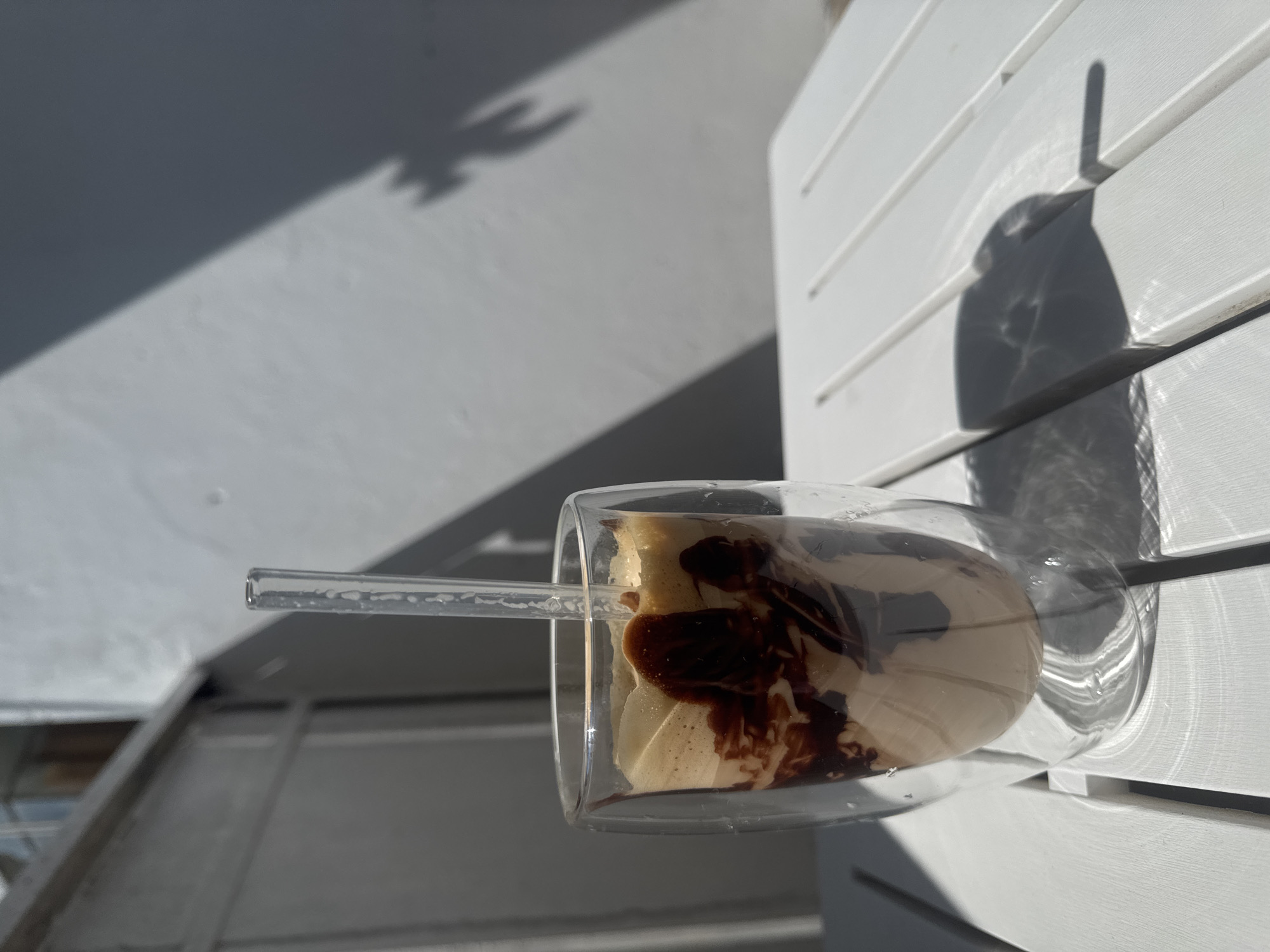 A glass of iced coffee with chocolate syrup is on a white slatted table outdoors. The coffee is in a double-walled glass, and a clear straw is inserted into it. The table is casting shadows from the sunlight. In the background, a white wall is visible, also with shadows on it, including what appears to be a plant shadow. The scene is likely on a balcony or patio in Varna, Bulgaria, given the style of architecture and the bright sunlight. There are no people or animals in the image.