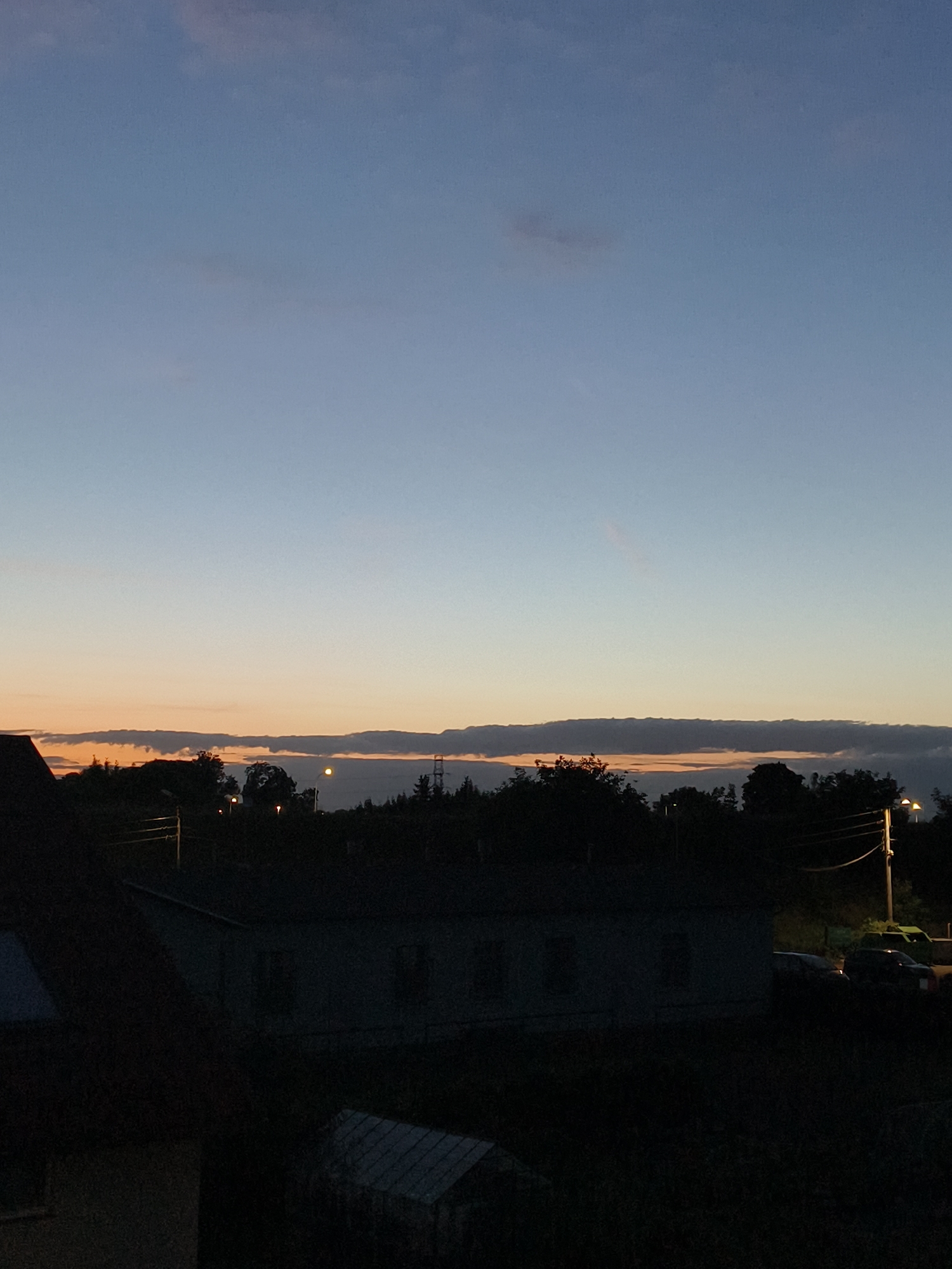 The image captures a rural or suburban landscape at dusk or dawn, likely in Tczew, Poland. The sky transitions from a pale blue at the top to hues of orange and yellow near the horizon, indicating the setting or rising sun. Long, horizontal clouds are visible against the warmer sky. Silhouetted against this backdrop are dark trees and what appears to be a cluster of buildings, possibly a small industrial or residential area. There are also streetlights visible, casting a soft glow. In the foreground, a dark roofline of a building occupies the left side of the frame. The overall impression is one of quietude and twilight.