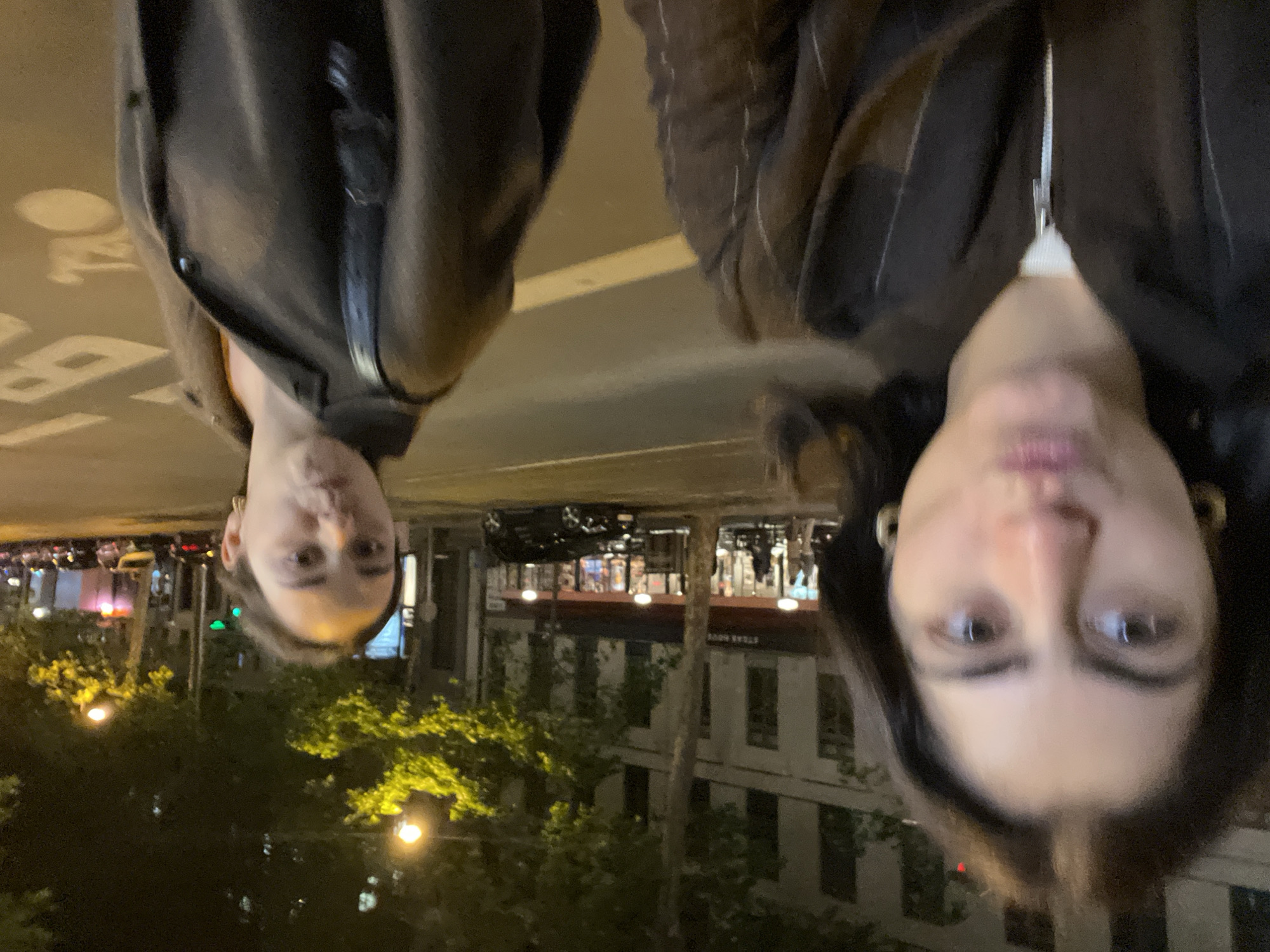 Two people are standing on a street at night in Paris, France. They are looking at the camera, with the person on the right closer to the camera. Buildings line the street and trees are in the foreground. The street has white lines and the trees are lit with street lights.