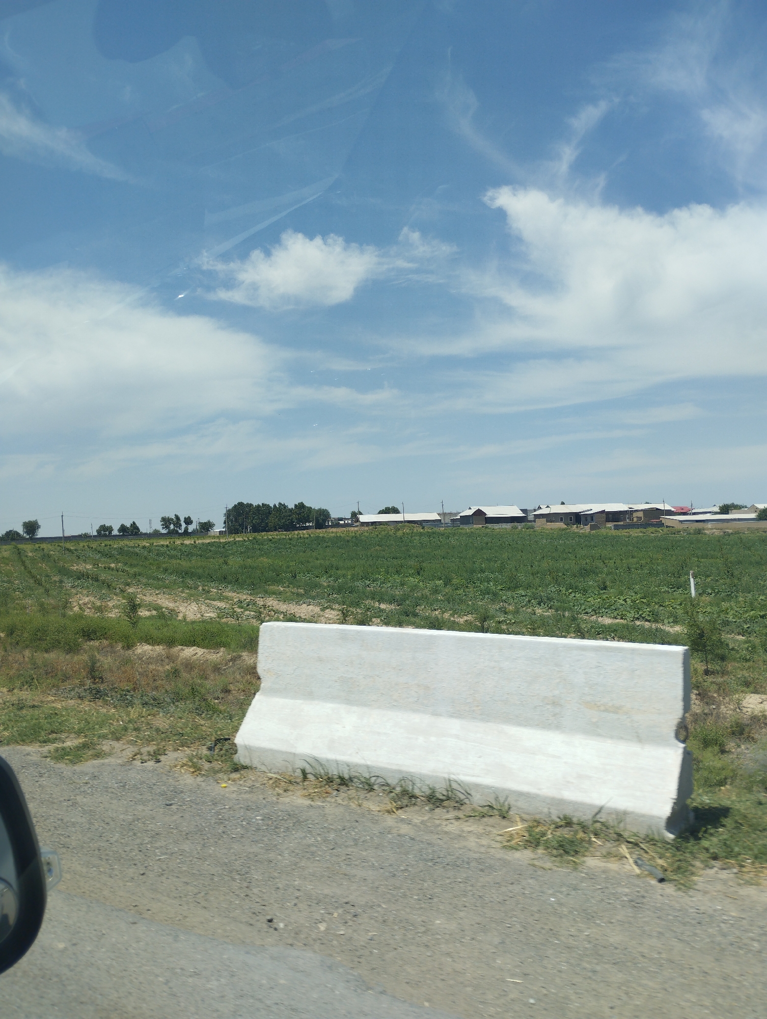 The image depicts a roadside view in Turkkishlak, Uzbekistan. In the foreground, a grey concrete barrier is positioned on the right side of the frame. Beyond the barrier, a dirt road and a field of green crops extend towards a line of buildings in the middle ground. The buildings appear to be residential or agricultural structures with white roofs, set against a backdrop of trees and a blue sky with scattered clouds. The overall scene suggests a rural or semi-rural environment.