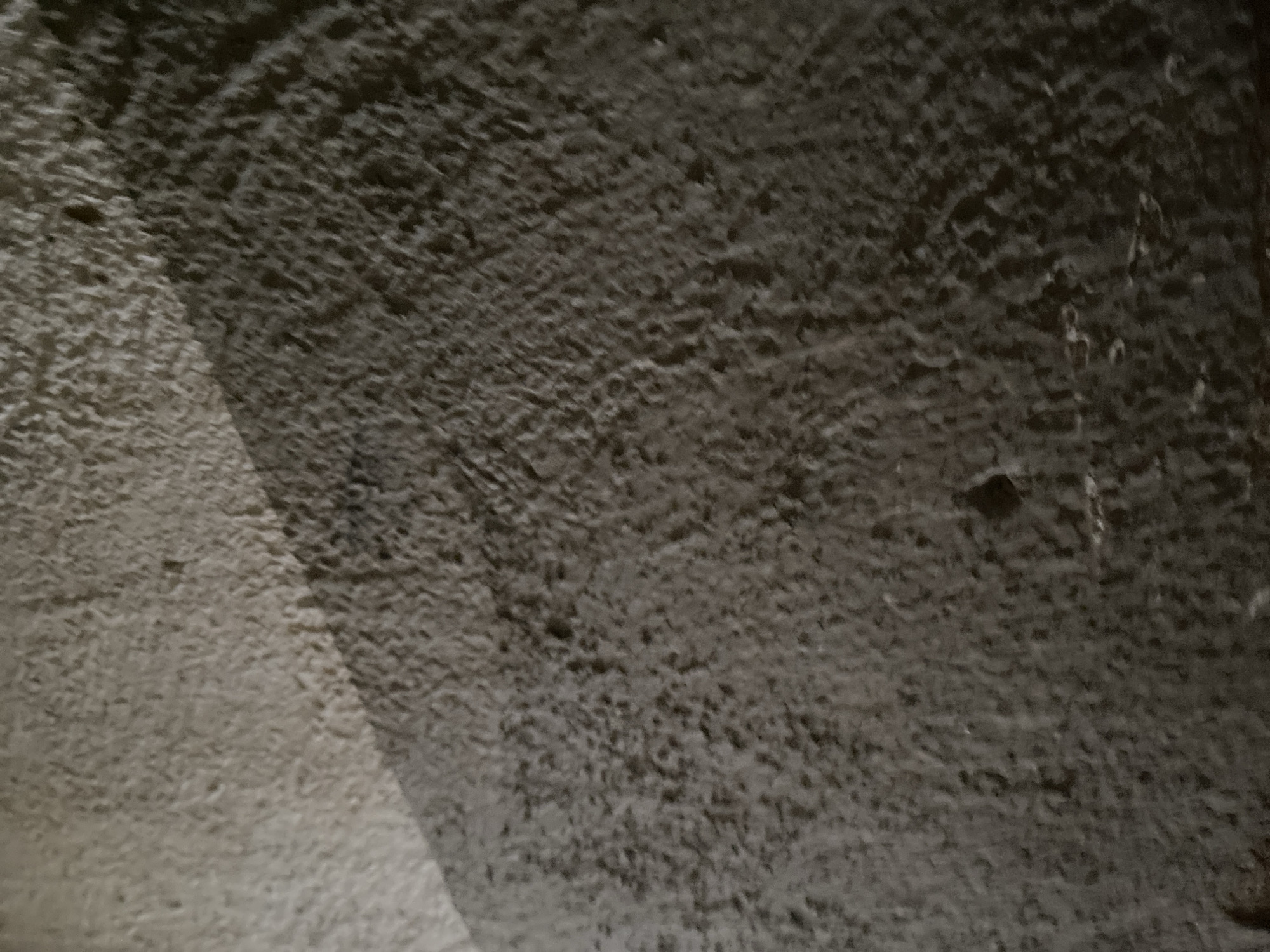 The image shows a close-up view of a rough, textured surface, likely a wall or ground. The surface is predominantly gray with variations in shade, indicating unevenness and possibly dampness or dirt. A distinct shadow cuts diagonally across the scene from the upper left to the lower right, suggesting an external light source. The texture appears granular and uneven, with small pits and bumps visible. There are no people or discernible objects in the image. The scene is likely located in Jauro Yinu, Nigeria, but specific landmarks or identifying features are absent. The focus is solely on the texture and light play on the surface.