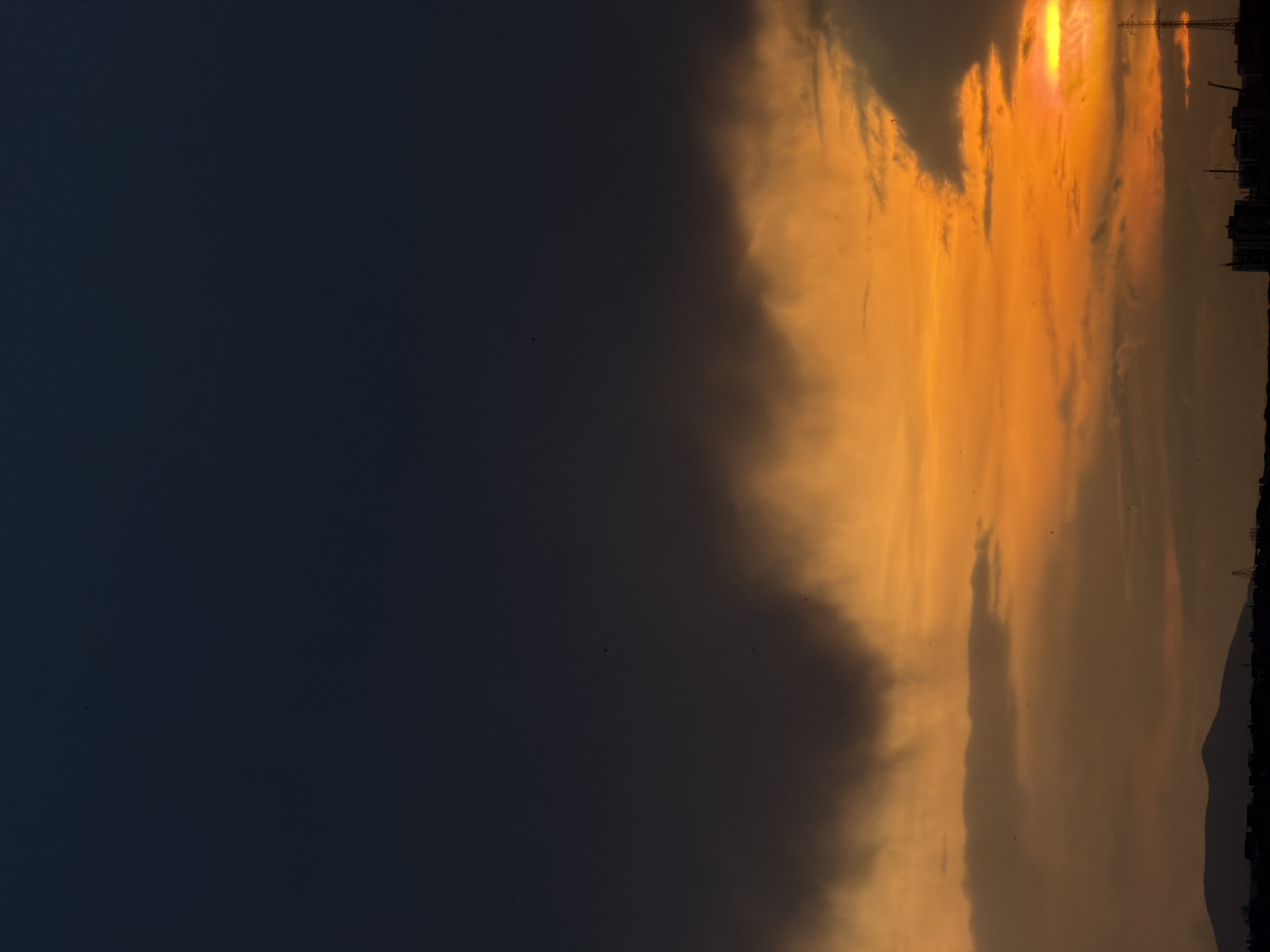 The image captures a vertical view of a twilight sky in Tbilisi, Georgia. The sky is divided into two distinct regions: the left side is a dark, navy blue, transitioning into a vibrant golden-orange sunset on the right. The sunset area features layered clouds, creating a gradient of light and shadow. In the background, a dark, silhouetted skyline of buildings and mountains is visible. Overhead power lines are also silhouetted against the bright sky. Small, dark bird silhouettes dot the sky.