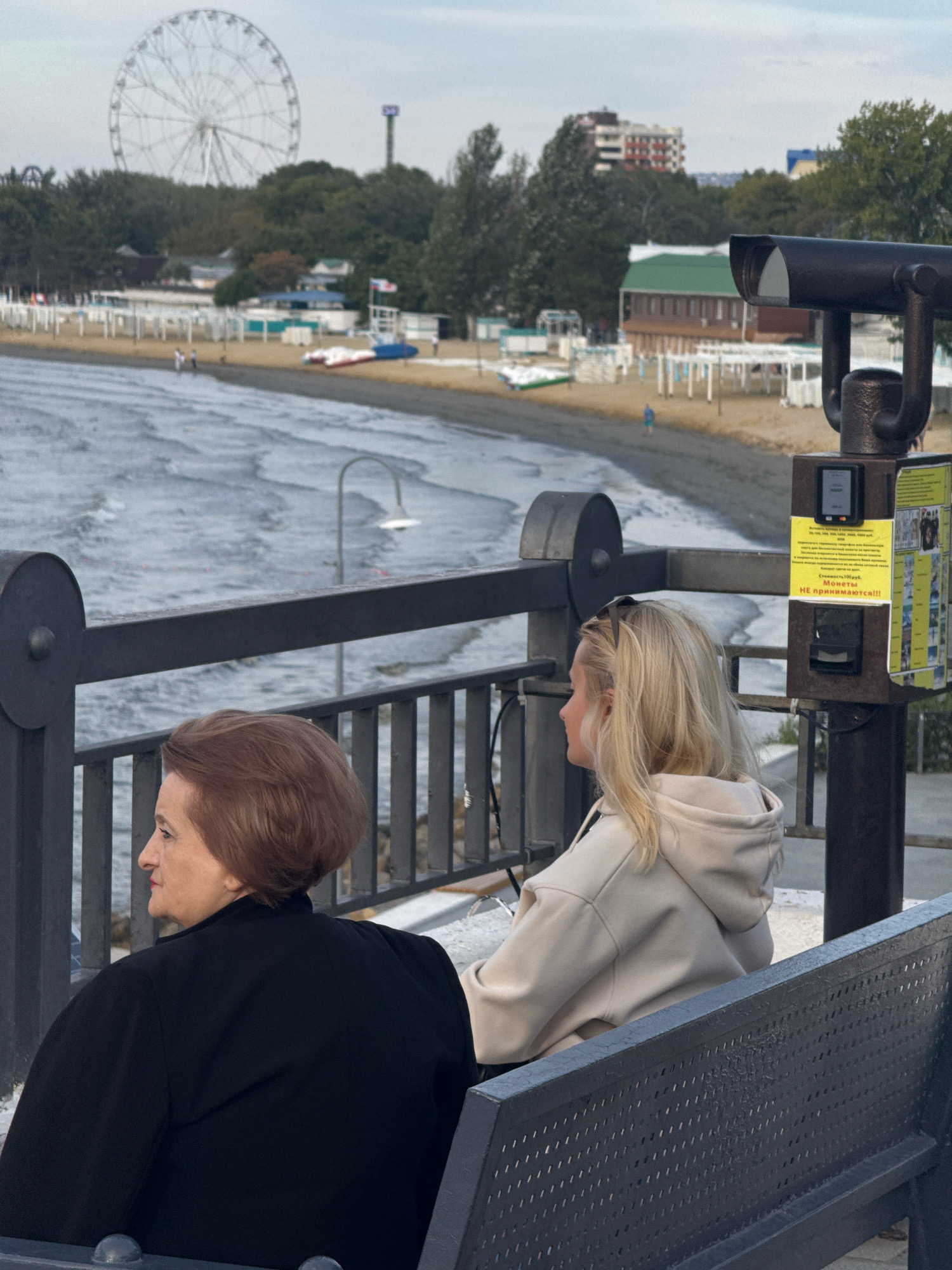 The image depicts an outdoor scene at a promenade overlooking the sea, likely in Anapa, Russia. Two women are seated on a dark metal bench, facing away from the camera. The woman on the left has short, auburn hair and is wearing a black jacket. The woman on the right has long, blonde hair and is wearing a beige hooded sweatshirt. In the foreground, a dark metal railing separates the bench area from the promenade. A coin-operated viewing scope is mounted on a post to the right of the bench. In the middle ground, a sandy beach is visible, with a few beach umbrellas and what appear to be beach chairs. The sea is visible in the background, with moderate waves. Further in the distance, a Ferris wheel, a tall tower with a light on top, and various buildings are visible, suggesting an urban or resort area. The sky is overcast.