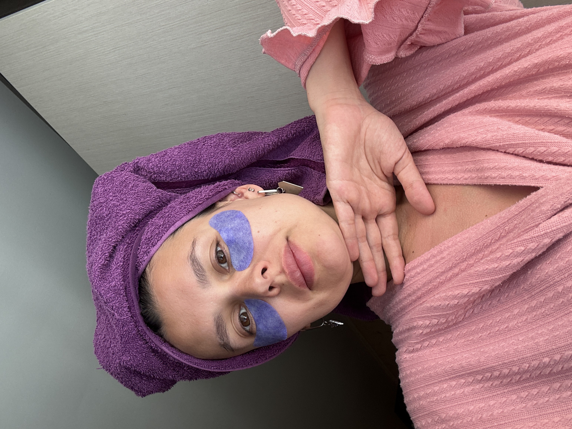 A woman is facing the camera, wearing blue eye patches and a purple towel on her head. She is wearing a pink sweater. The background is gray. The location is Chaikovsky, Russia.