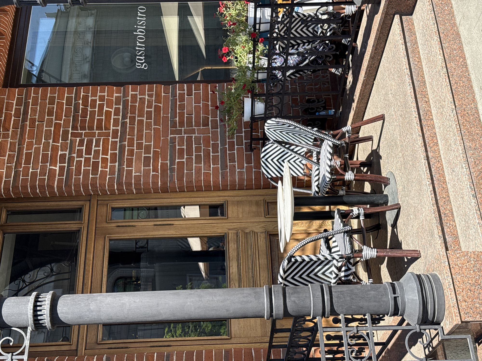 The image depicts an outdoor seating area of a restaurant with a brick facade in Krasnoyarsk, Russia. A tall, grey cylindrical lamp post stands to the left, positioned in front of a wooden door and window. In the center, a round white table is set with two chairs, each featuring a black and white zigzag pattern. These chairs have dark wooden legs. Behind the table and chairs, a black metal railing encloses the seating area. Beyond the railing, several white pots containing red flowers are displayed on a ledge against the brick wall. To the right, a window displays the word "gastro bistro" in white lettering, suggesting the establishment's name. The ground is paved with a textured material, and granite steps lead up to the seating area. The lighting suggests it is a sunny day. There are no people visible in the image.