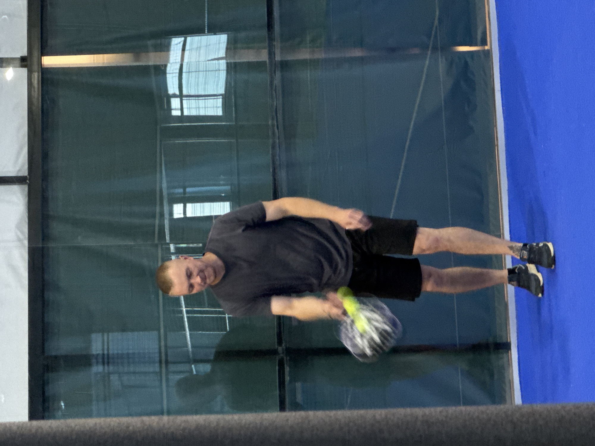 The media file shows a man playing a sport, likely padel or a similar racket sport, indoors. He is wearing a dark t-shirt and dark shorts, and is holding a racket with a fluorescent yellow ball. The man appears to be in motion, possibly in the middle of hitting the ball, as the racket and ball are blurred. He is on a blue court with glass walls, suggesting an indoor sports facility. The lighting appears to be artificial, typical of an indoor environment. The man has fair skin and short, light brown hair. His expression is neutral, focused on the game. The background shows reflections on the glass walls, possibly of other players or the facility's structure. No text is visible in the image. The overall scene depicts an active moment during a sports game.