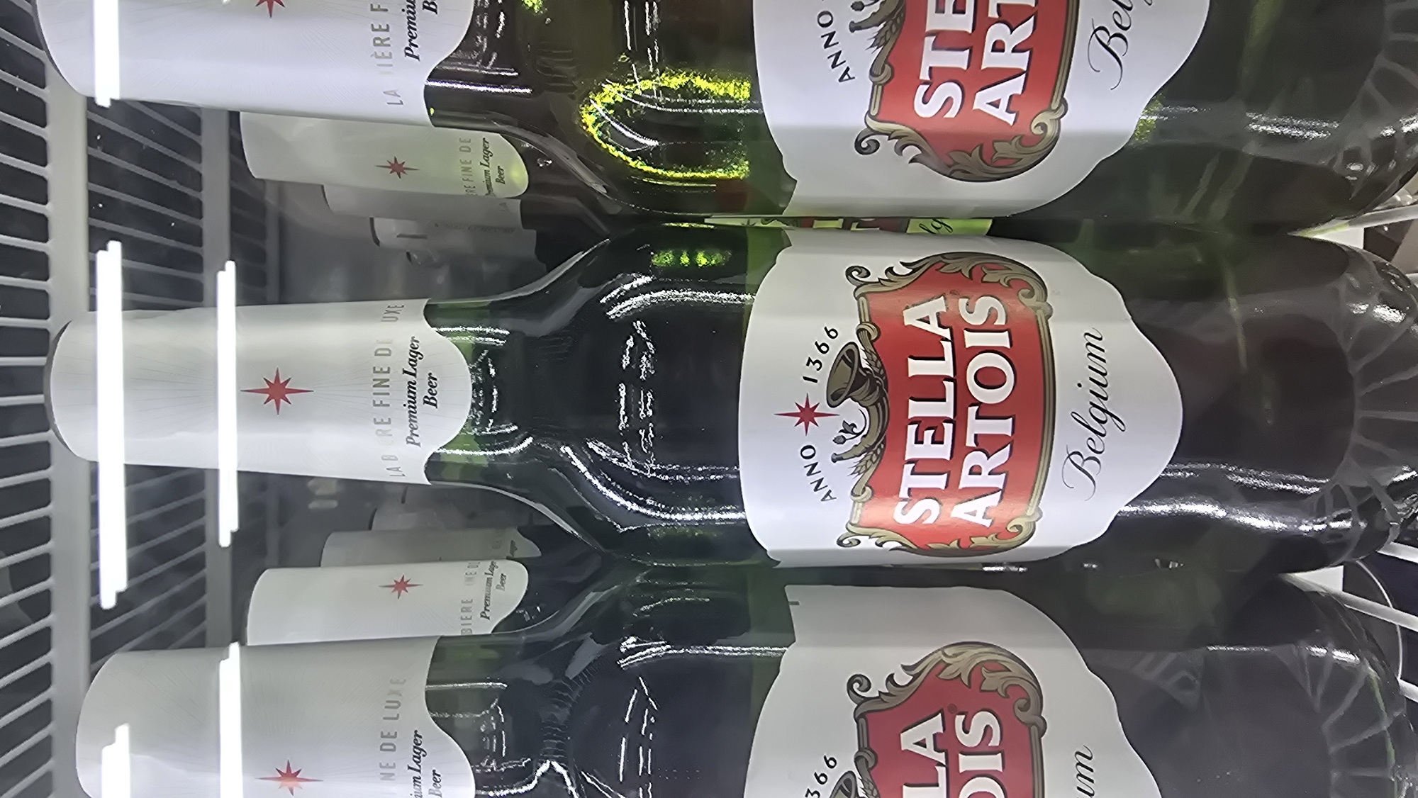 The image shows a close-up view of several bottles of Stella Artois beer in a refrigerator. The bottles are green glass with white labels that feature the Stella Artois logo, including the text "STELLA ARTOIS" and "Belgium". Some of the labels also show "Anno 1366" and a red star. The lighting in the image appears to be from fluorescent lights, possibly within a store or a bar. There are no people visible in the image. The background is blurred and appears to be the interior of a refrigerator with shelving. The scene is generic and does not provide any specific information about its location in Moscow, Russia.