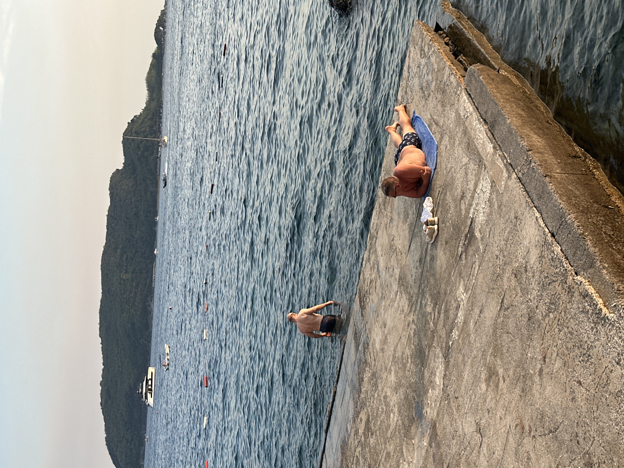 The image shows a concrete pier extending into the sea in Budva, Montenegro. Two men are visible. One man is standing in the shallow water at the edge of the pier, facing away from the camera. He is wearing swim trunks. The other man is lying on a blue towel on the pier, facing towards the water. He is wearing swim trunks and sunglasses. There are a pair of shoes and a white towel next to him. In the background, there is a large body of water with a distant green, tree-covered island. Several buoys are floating in the water closer to the shore. A large white yacht is visible in the distance, as well as other smaller boats. The sky is clear and the sun appears to be setting, casting a warm glow on the scene. The pier is cracked and weathered.
