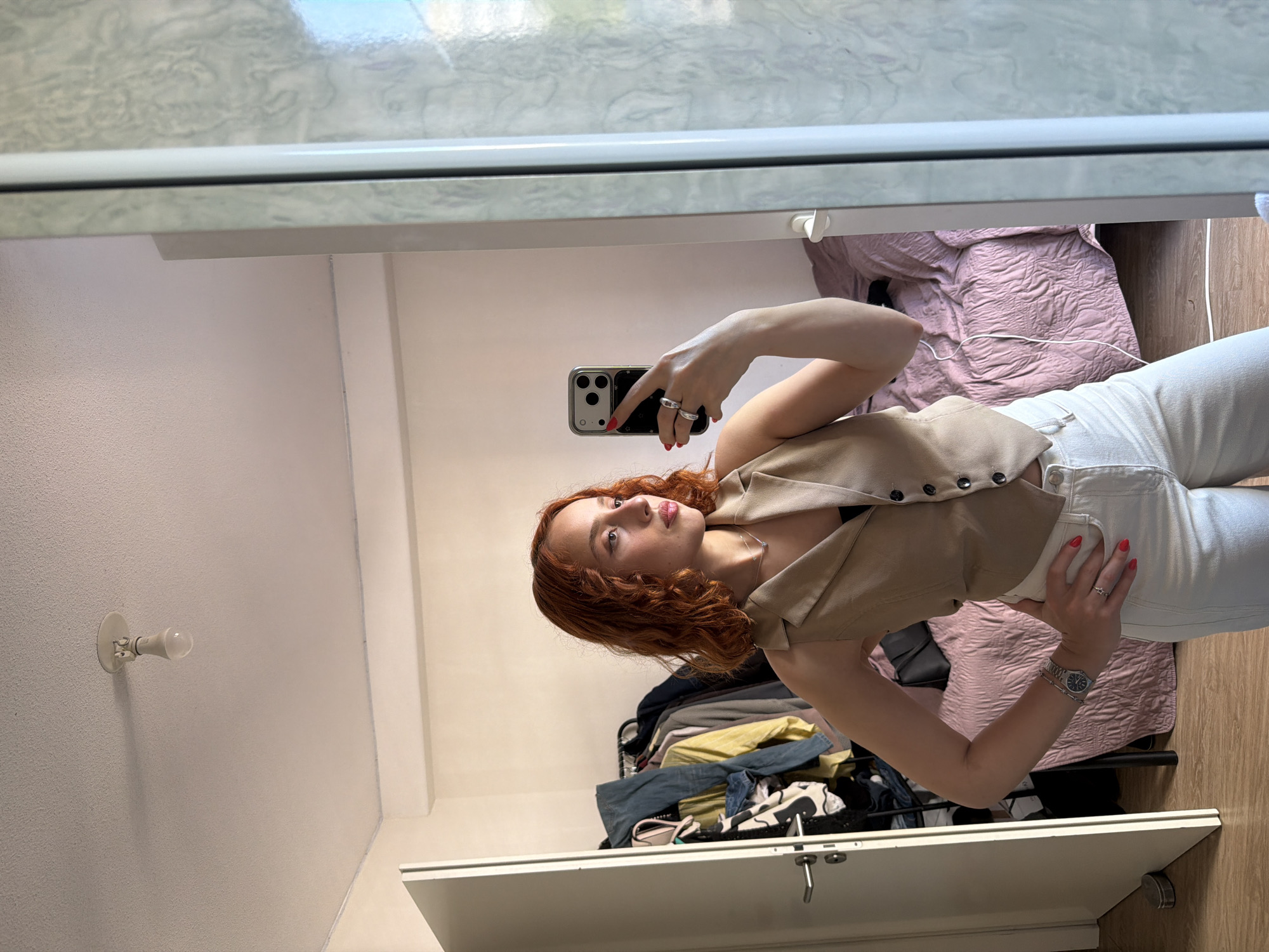 The image depicts a woman with red, curly hair taking a mirror selfie in what appears to be a bedroom. She is wearing a beige vest over a dark top and white pants. Her right hand is holding a smartphone, and her left hand is on her hip. There is a clothing rack with various garments behind her, a bed with a pink textured comforter, and a door to her left. The environment appears to be indoors, with a light fixture visible on the ceiling. The overall lighting suggests daytime. There is no indication of the location being in Strasbourg, France.