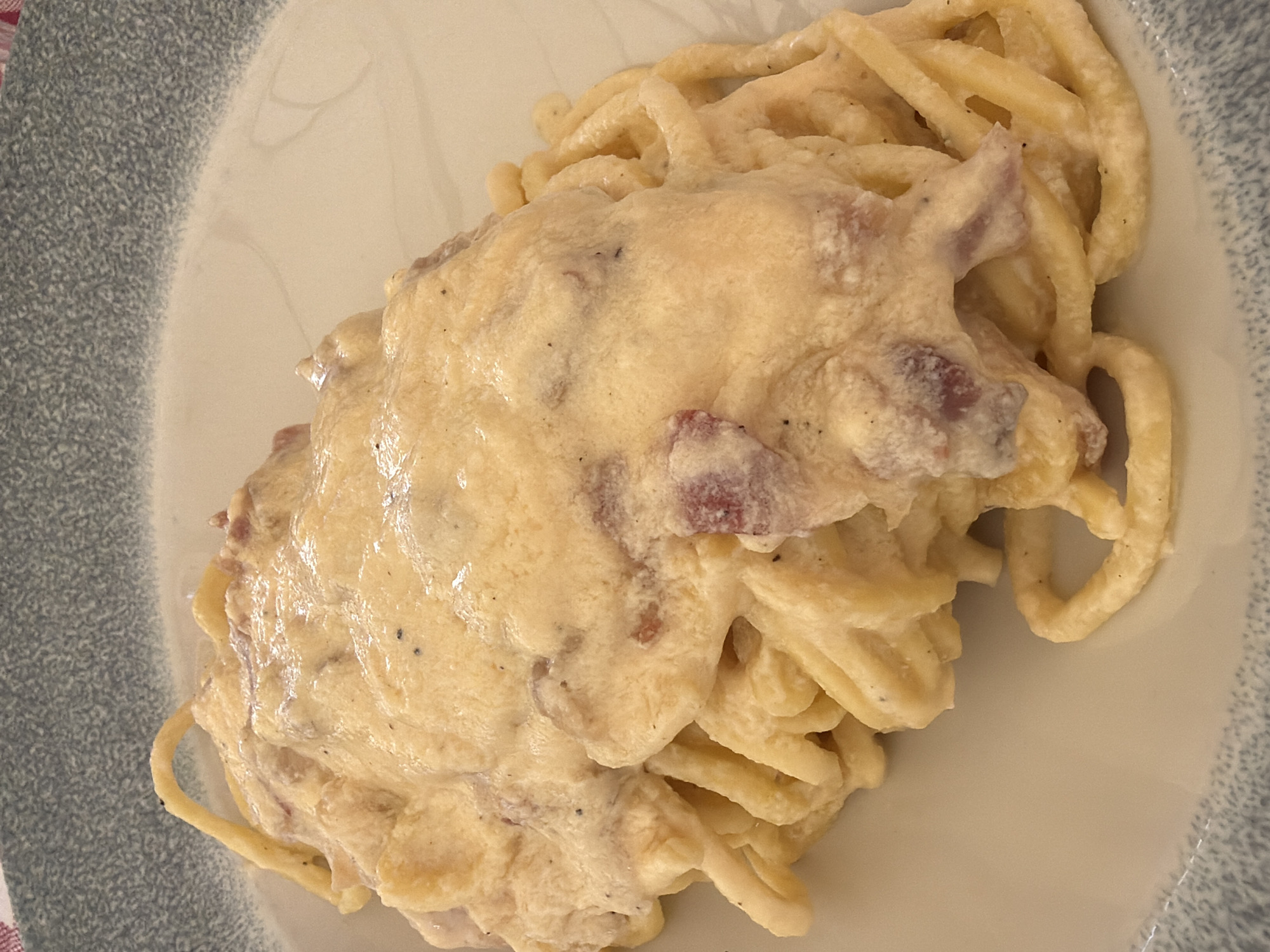 A plate of spaghetti carbonara is centered in the image. The pasta is coated in a thick, creamy sauce with visible pieces of cured pork. The plate is white with a grey and white textured border. The location, Como, Italy, is not inferable from the image.