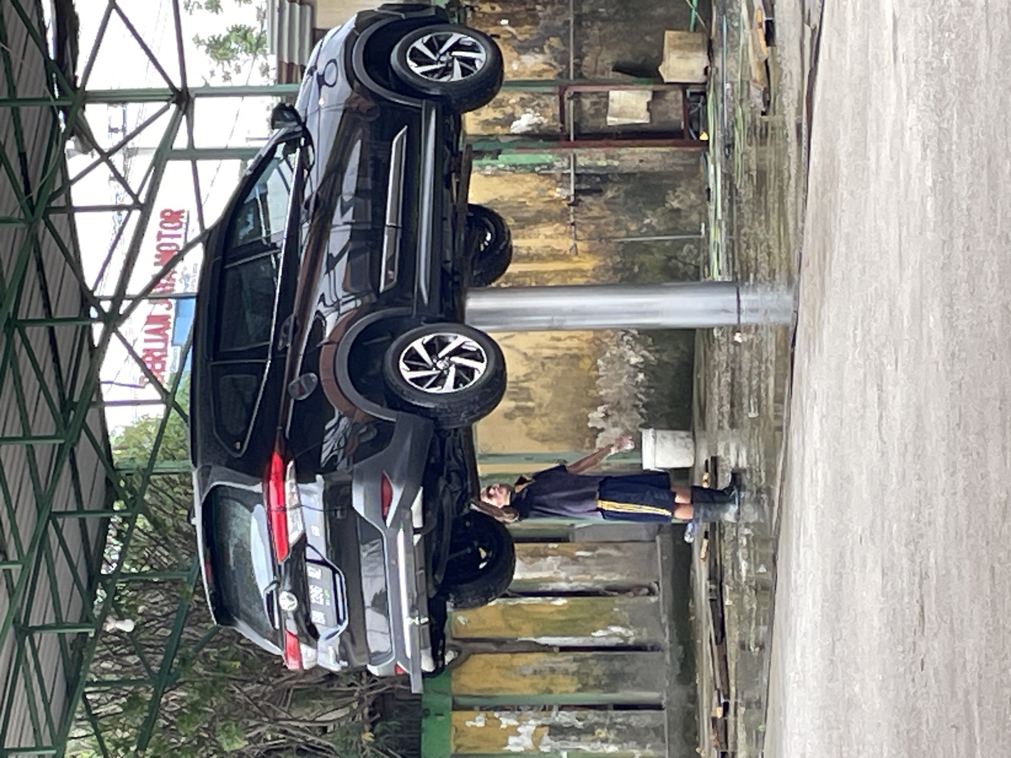 A black SUV is elevated on a car lift at an outdoor car wash in West Sidomulyo Subdistrict, Indonesia. A male attendant, wearing a dark blue t-shirt with yellow trim, blue shorts with yellow trim, and black boots, stands beneath the car, holding his right arm up towards the underside of the vehicle. He appears to be cleaning or inspecting the car. The car wash area is wet, with standing water on the ground. In the background, there is a building with a sign that reads "PERLIAN JAYA MOTOR". Green metal framing supports a roof structure over the car lift. Trees and vegetation are visible behind the building.