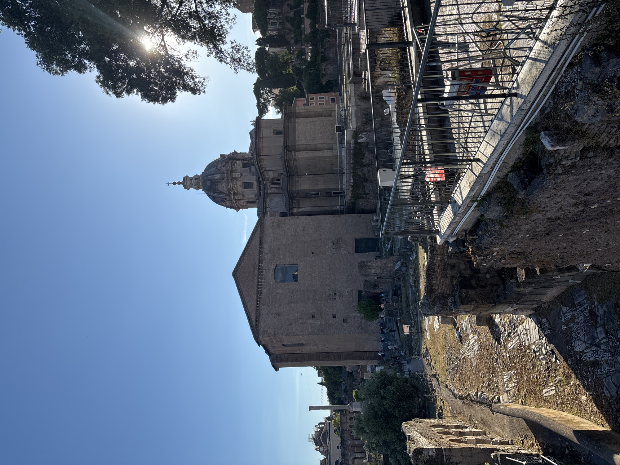 This image depicts a scene in Rome, Italy, likely within or overlooking the Roman Forum. The foreground includes remnants of ancient structures, showing weathered stone and archaeological remains. A metal railing and modern walkway are present, suggesting a designated tourist viewpoint. In the midground, a large brick building with a prominent dome dominates the view, appearing to be a church or similar historical edifice. The sky is blue, with the sun visible through the leaves of a tree in the upper-left corner. The environment is sunny and outdoors, suggesting a daytime setting. There are a few people visible, adding a sense of human activity to the historical site.