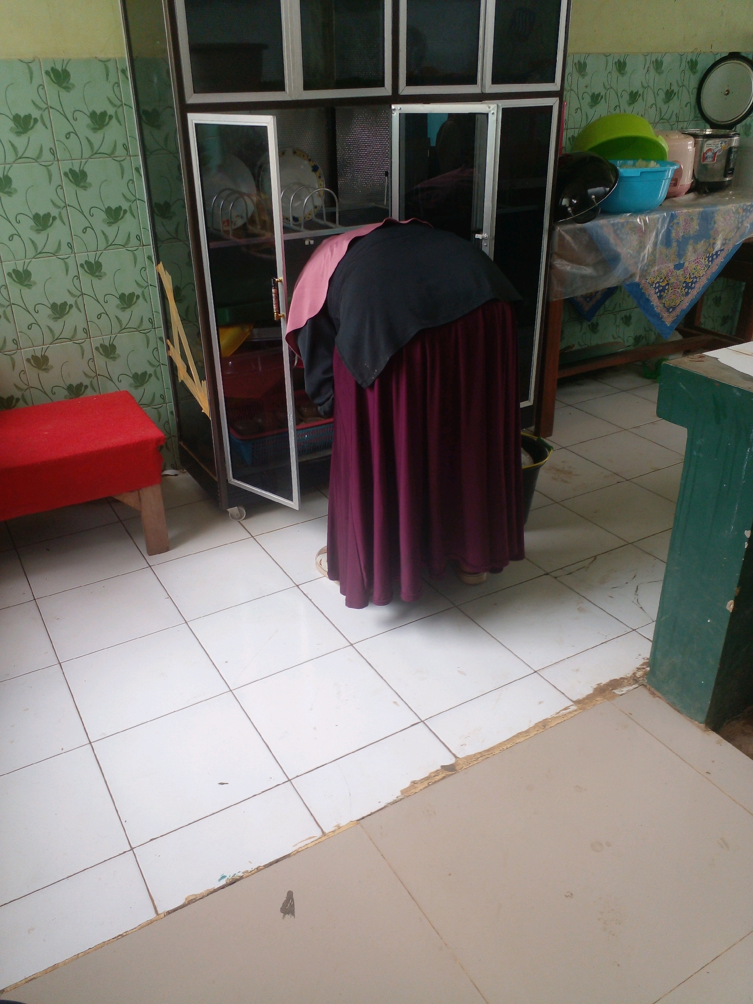 A person wearing a dark blue hijab and a maroon skirt is bending over in front of a glass-fronted cabinet in a room with tiled floors and green floral patterned tiles on the wall. The cabinet contains dishes and various kitchen items. To the left of the cabinet is a small red-covered table. To the right of the cabinet, on a table covered with a patterned cloth, are several plastic bowls and a rice cooker. The overall scene depicts a domestic environment within a building.
