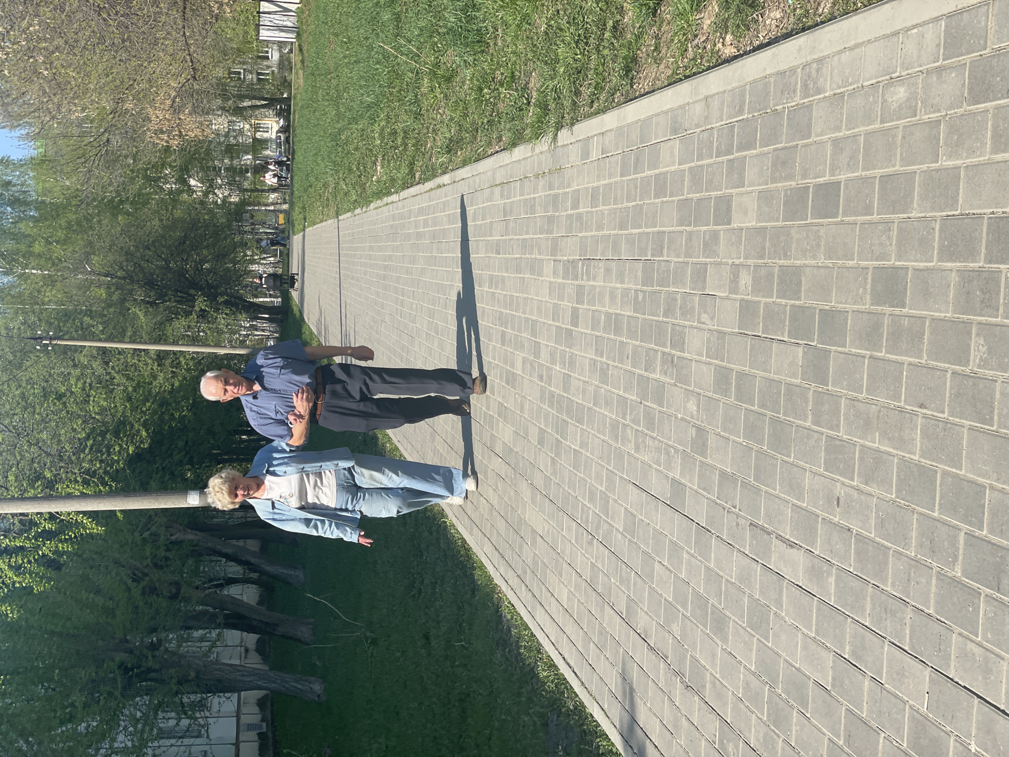 The image shows two elderly people walking on a brick paved path next to a grassy area with trees in Izhevsk, Russia. The man is wearing a blue shirt and dark pants, while the woman is wearing a white shirt with a light blue jacket and jeans. Their shadows are cast on the path. The path is made of gray bricks and is bordered by grass. Trees and buildings are visible in the background.