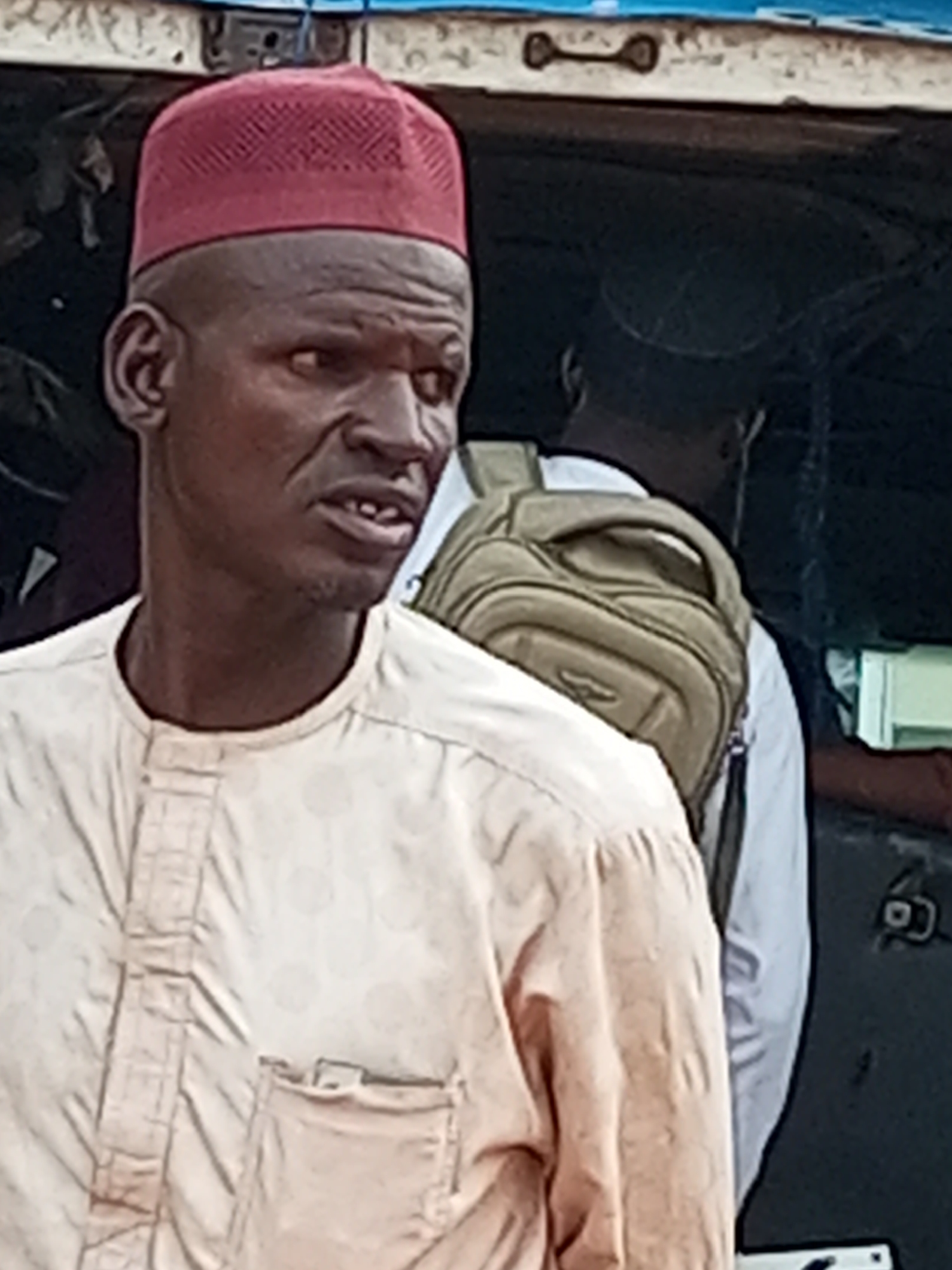 In Gombe, Nigeria, a man wearing a red kufi and a light-colored, buttoned shirt stands in front of a dark background, possibly a vehicle. His eyes are open, and his mouth is slightly ajar, revealing some teeth. Another person with a backpack is visible behind him. The overall lighting suggests an outdoor setting.
