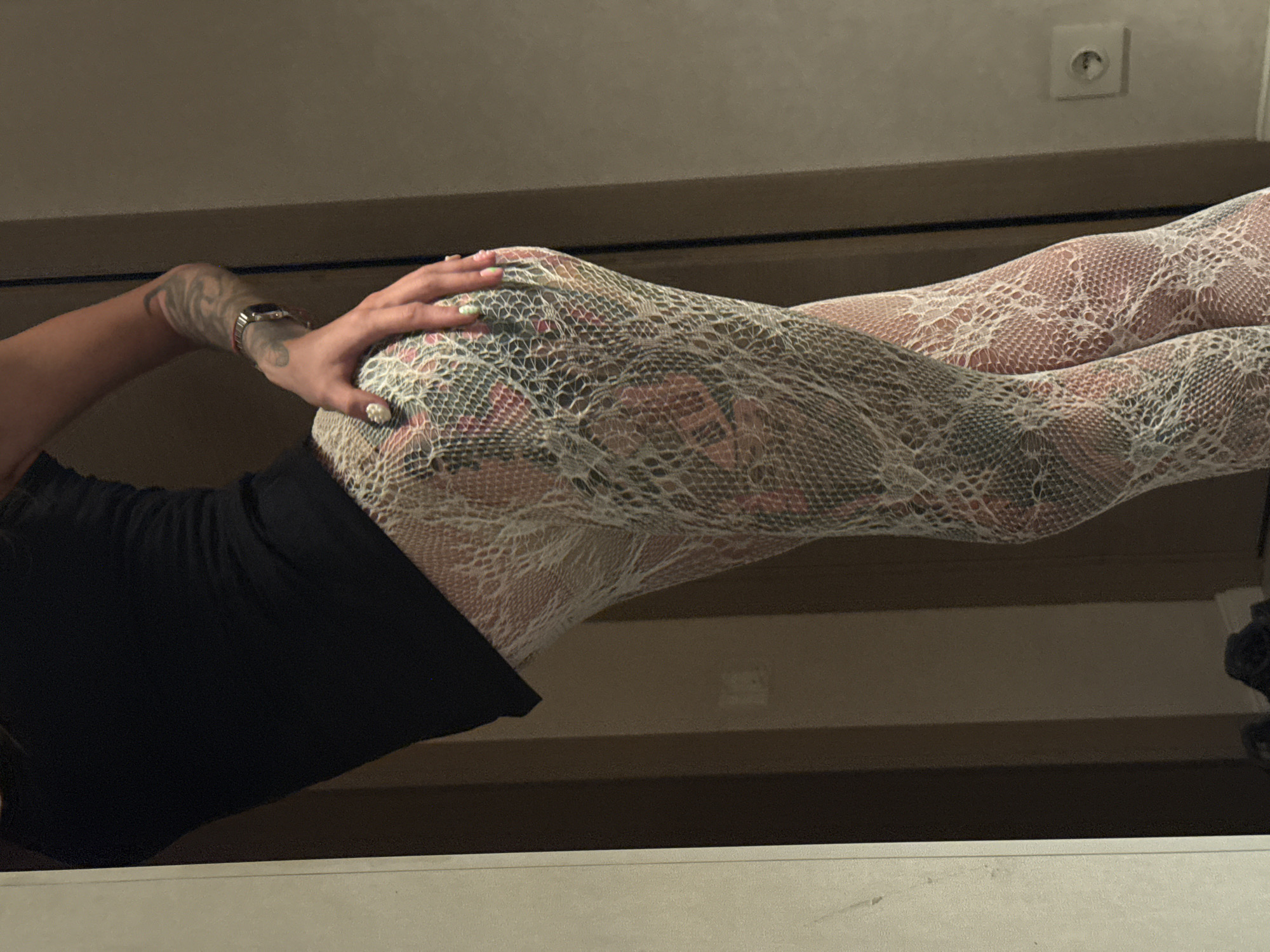 The image shows a person from the waist down, wearing a black tank top and white lace patterned tights with a snake print visible underneath. The person is leaning against a wall, with their hand resting on their hip. The background appears to be an indoor setting with a wall and a doorway. There is an electrical outlet on the wall. The image does not provide any information to determine the location in Moscow, Russia.