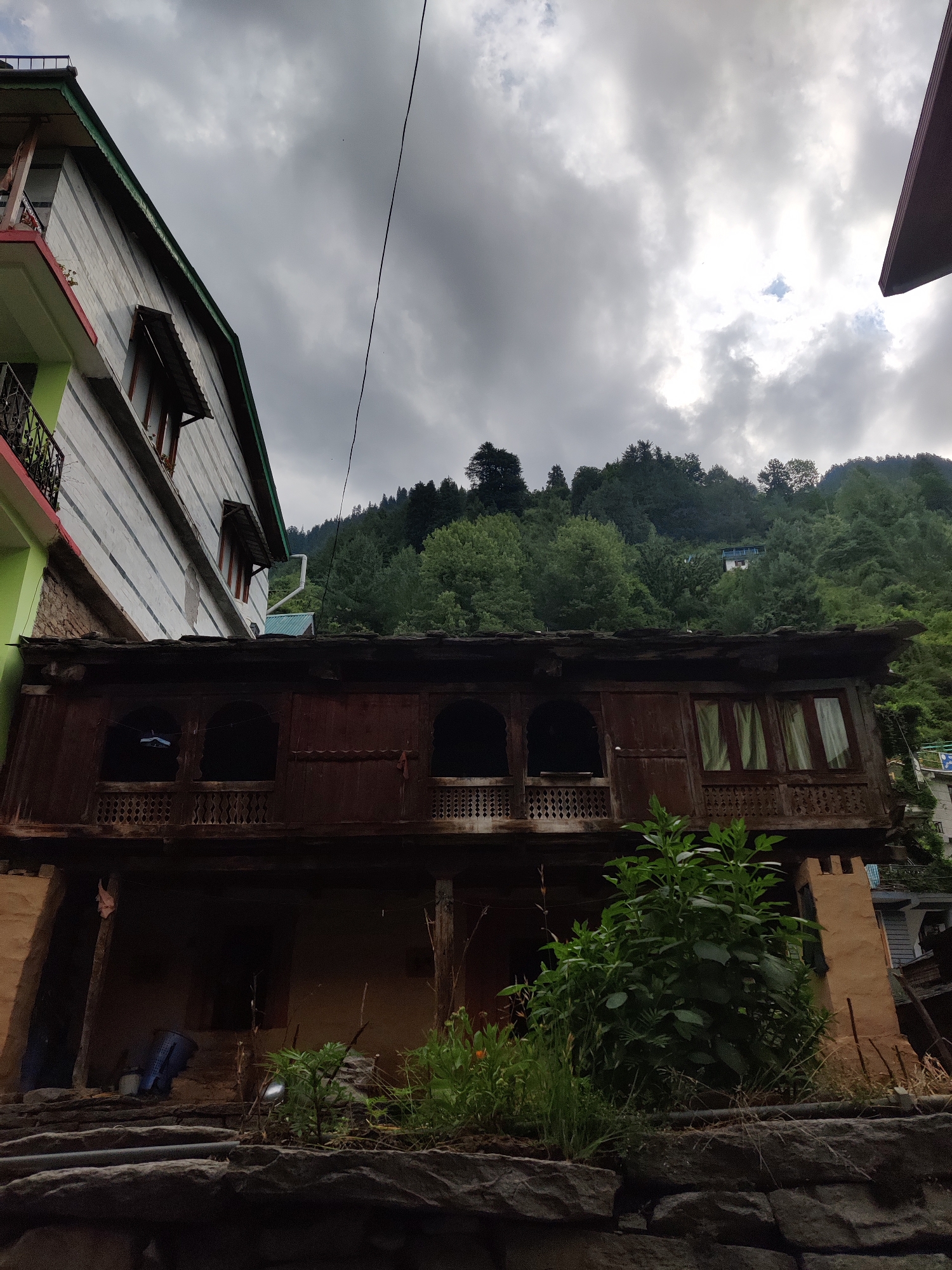 A low-angle, wide shot captures a view of traditional wooden architecture in Vashisht, India, under a cloudy sky. The foreground features a stone wall and stairs leading up to a multi-story building. The building on the left has a modern facade with white siding and horizontal grey stripes, accented with a green and red trim on the upper balcony. Attached to this building, a more traditional, dark wooden structure juts out. This wooden structure has an elaborately carved balustrade along its upper floor, with arched window openings and curtained windows on the right side. Below this wooden section, an open, dimly lit area appears to be an entrance or storage space, with a blue bucket visible. Lush green foliage and some flowering plants are growing in front of this lower level. In the background, a dense forest of green trees covers a steep hillside, with a small white building visible higher up. A black power line stretches diagonally across the frame from the upper left. The overall atmosphere is overcast, suggesting a possibly rainy or humid day. No people are visible in the scene.
