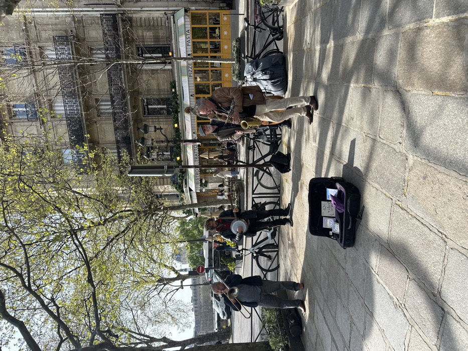 An outdoor scene depicts a jazz quartet performing on a stone-paved sidewalk in Paris, France, near the Seine River. The location is identified as the area opposite the Institut de France, specifically near the building housing "Le Carrousel" bistro (visible signage: "BISTRO TRATTORIA PIZZA PASTA" with yellow paneling and a green-and-white striped awning) at 11 Quai Anatole France, 75007 Paris. The distant dome of the Institut de France is visible across the river.
Three musicians are actively playing instruments, and a fourth is present with an upright bass. From left to right: a male in a dark jacket and grey trousers plays a trombone; a male with a beard and curly hair, wearing a dark jacket and red scarf, plays a banjo; a male in a brown jacket and flat cap stands behind the saxophonist, partially obscured, with what appears to be an upright bass; and a male with short grey hair and a beard, in a light brown jacket and light trousers, plays a tenor saxophone.
In the immediate foreground, an open black instrument case rests on the paving stones, containing several CDs, money, and a sign displaying "10€" along with other text, indicating a street performance setup for donations and sales. A purple cloth item is also inside the case.
Behind the musicians, several bicycles are parked against a metal street barrier, including a white e-bike and a black road bike. A large, dark grey bag or instrument case is on the ground near the rightmost bicycle. The street behind the barrier shows parked vehicles and a red circular no-entry sign. Beyond the street is a multi-story building featuring classical Haussmannian architecture with balconies, housing the restaurant on the ground floor. Trees with nascent green foliage line the street. The scene is illuminated by bright sunlight, casting distinct shadows on the paving stones.