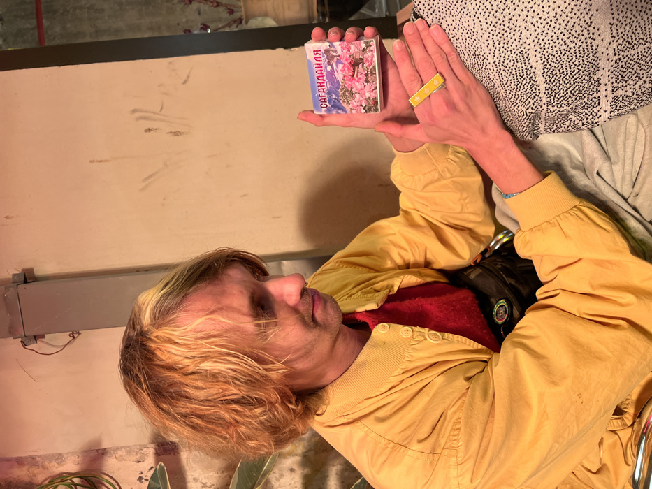 A male individual with wavy, shoulder-length, reddish-blonde hair and a silver nose ring is depicted seated indoors. He wears a yellow bomber jacket over a red sweater and a dark fanny pack around his waist. In his right hand, he holds a small, white rectangular package featuring an image of snow-capped mountains and pink flowers, with the Cyrillic text "САГАНДАЙЛЯ" in red at the top. His left hand is open, displaying a yellow rectangular object adorned with white stars resting on his fingers. His gaze is directed downwards towards the objects. The background consists of a light beige wall with faint marks. A gray metal conduit with wires is affixed to the upper left wall, adjacent to a green-leafed plant. To the right, a cream-colored vase holds dried botanical elements. The specific location within Tel-Aviv, Israel, is not discernible from the image.