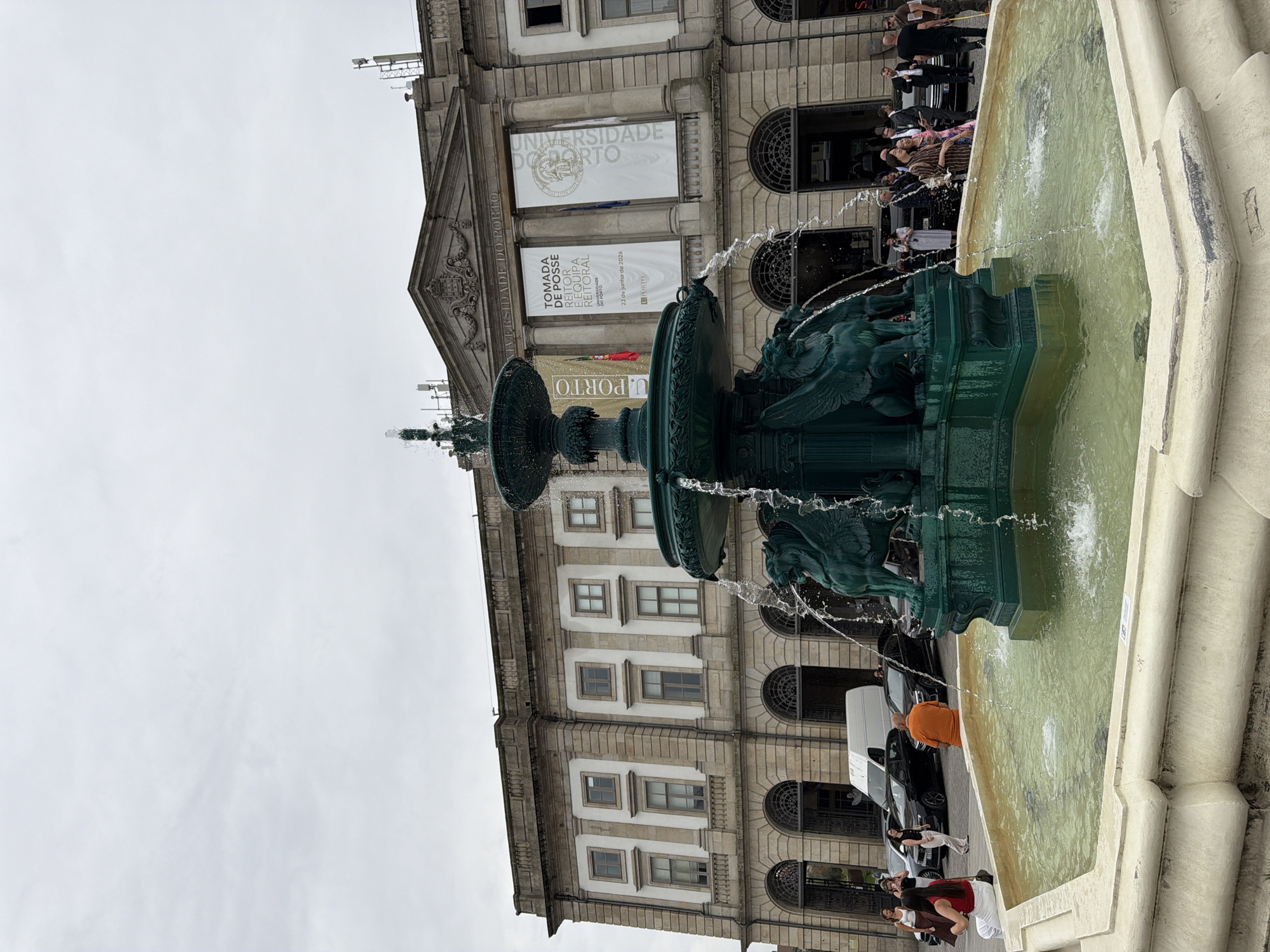 The image shows a wide shot of the Fountain of the Four Rivers (Fontes dos Quatro Rios) in front of the historic building of the Faculty of Sciences of the University of Porto, located in Porto, Portugal. The fountain, made of dark green metal, features multiple tiers and decorative sculptures of winged creatures. Water is actively spraying from the fountain into the basin, which is surrounded by a cream-colored stone border.
Several people are visible in the scene. In the foreground, to the left, a woman in a red top and white pants is sitting on the fountain's edge, facing away from the camera. To her right, a man in an orange t-shirt is sitting with his back to the camera, also by the fountain. Further back, a group of people are standing near the entrance of the building and by the fountain, some appearing to be tourists or attending an event.
Behind the fountain is the imposing, classical facade of the University of Porto building, constructed with stone and featuring arched doorways on the ground floor and a series of rectangular windows on the upper levels. Two large white banners are hung on the facade, one with text that appears to be in Portuguese, and the other with an emblem and text. Several cars are parked in the background, including a white van and several dark sedans. The sky is overcast, indicating a cloudy day.