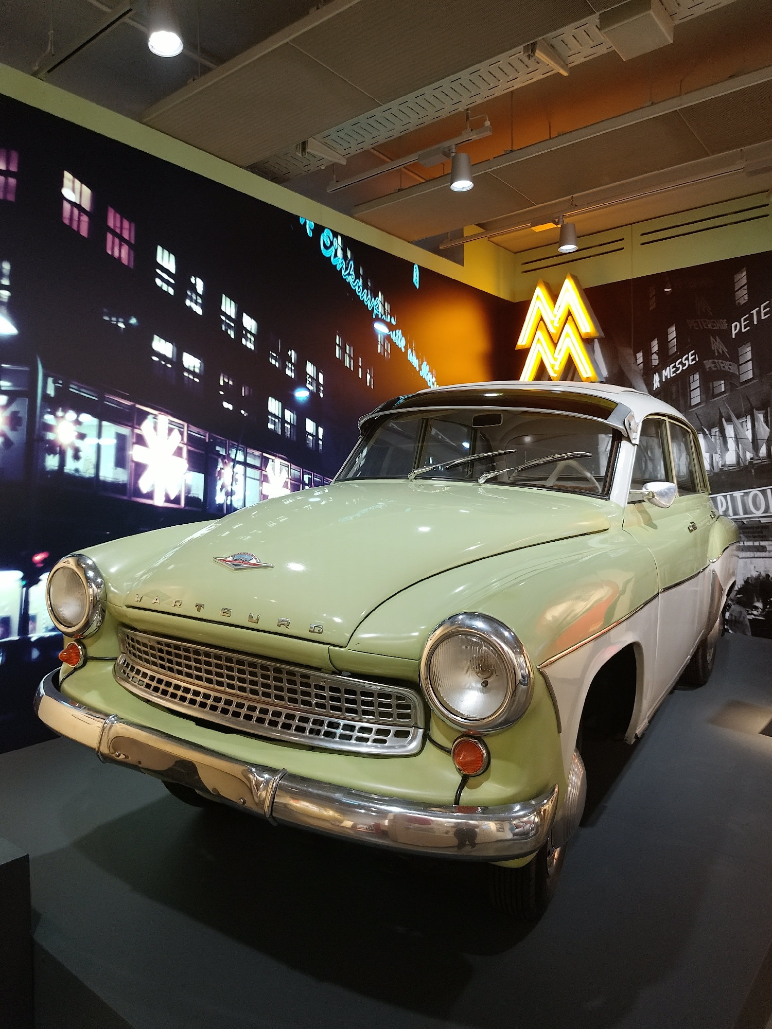 The image shows a two-tone green and white vintage Wartburg automobile displayed in a museum setting. The car is positioned in the foreground, facing the viewer at a slight angle. Behind it, a large wall mural depicts a cityscape at night, with illuminated buildings and a neon sign that reads "Messe" and "PETERHOF," suggesting a trade fair or exhibition location. An additional neon sign shaped like the letter "M" is visible above the car. The floor is dark gray, and the lighting in the space is provided by overhead track lighting.