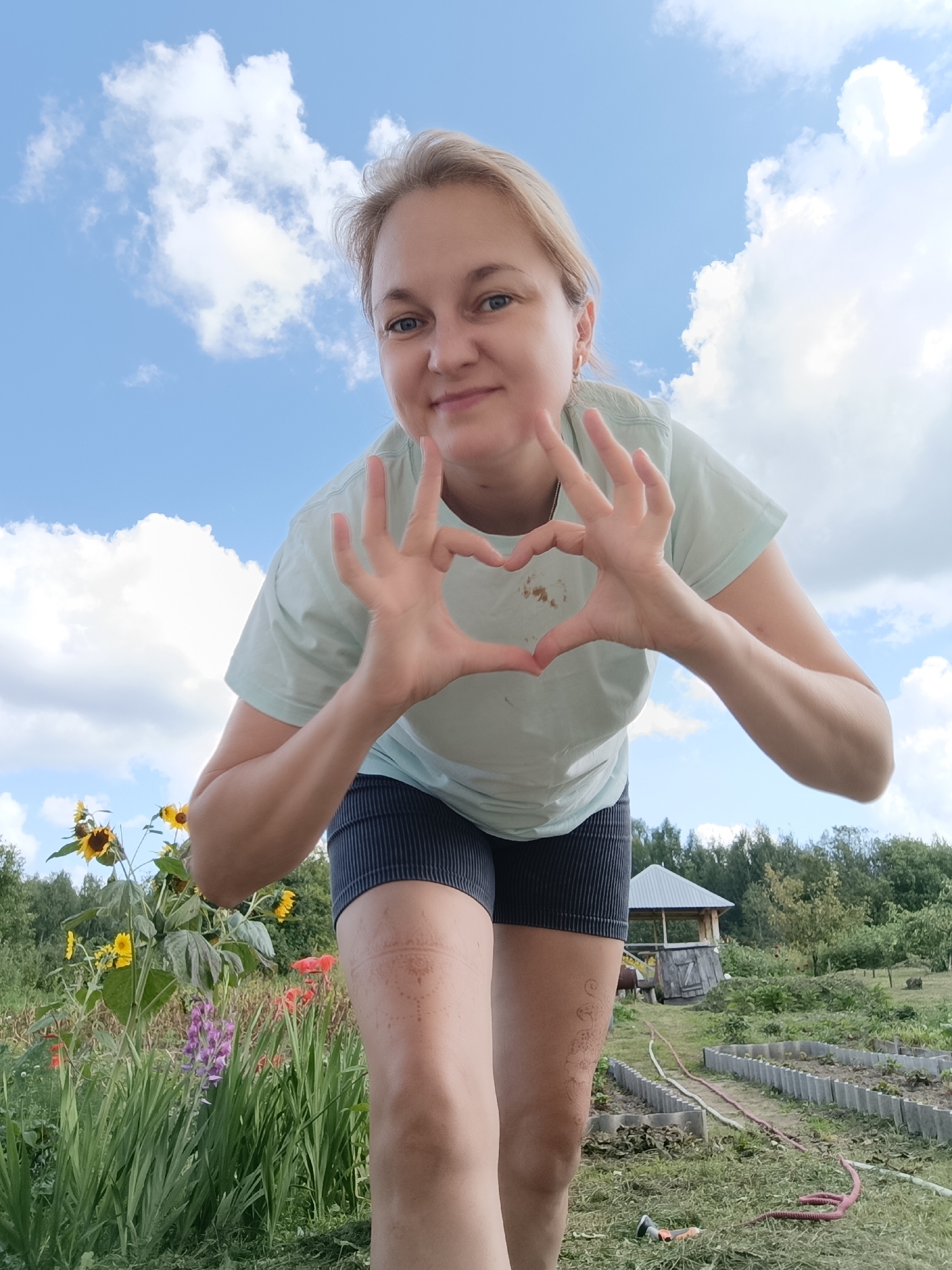 A blonde woman with blue eyes is photographed from a low angle, forming a heart shape with her hands in front of her chest. She is wearing a light teal t-shirt and dark striped shorts. Her expression is a warm smile, conveying a sense of joy and affection. Behind her, a vibrant garden scene unfolds under a partly cloudy blue sky. Tall sunflowers and other colorful flowers, including purple lupine and red gladioli, are in bloom, surrounded by lush green foliage. In the background, a rustic wooden gazebo with a grey roof stands amidst trees, suggesting a rural or garden setting. The ground is covered in grass, with some garden beds and hoses visible, indicating active gardening. A faint circular pattern is visible on the woman's right thigh, possibly a henna tattoo or temporary marking. The lighting suggests it is daytime, likely late morning or afternoon, with ample sunlight illuminating the scene. The overall atmosphere is cheerful and peaceful, capturing a moment of connection with nature. The location context provided is Dubovaya, Russia.