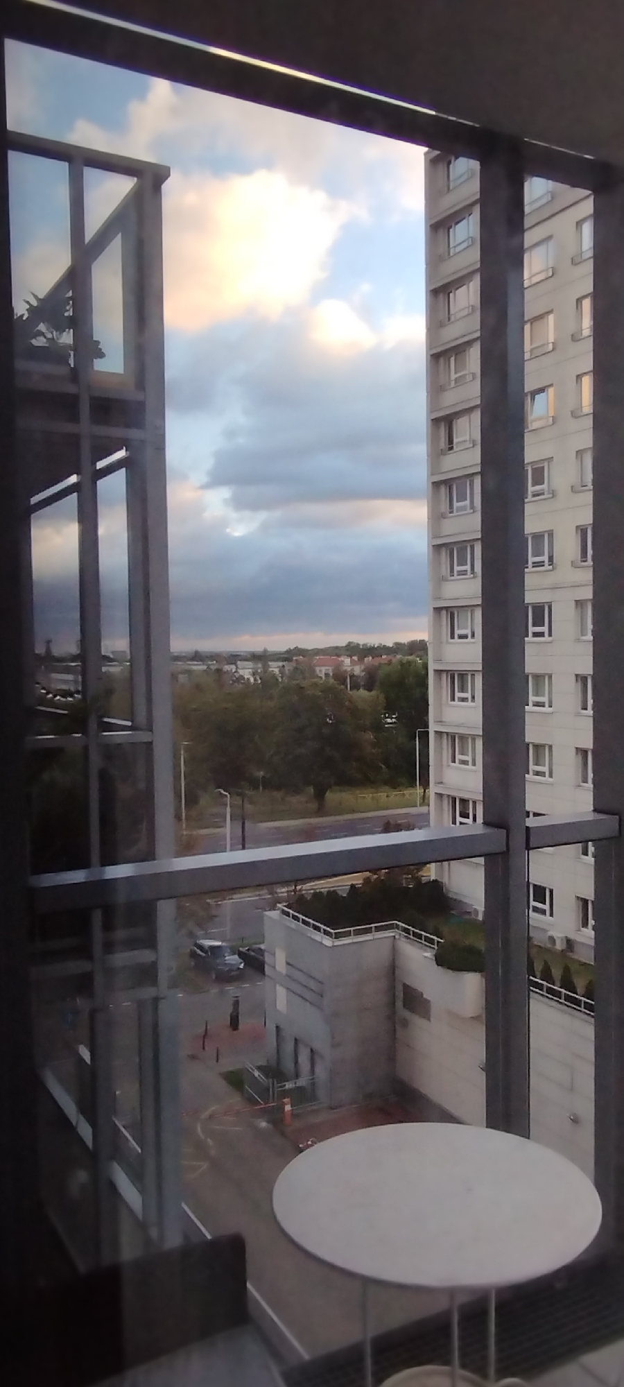The image shows a view from a balcony overlooking a street in Warsaw, Poland. In the foreground, a white round table with thin metal legs is partially visible. A dark chair is also visible to the left of the table. Beyond the balcony railing, a light-colored apartment building with multiple windows occupies the right side of the frame. The sky is cloudy with patches of light, suggesting either dawn or dusk. In the mid-ground, there are trees and some lower buildings, and a street with parked cars and construction barriers is visible. The scene appears to be a residential area within the city.