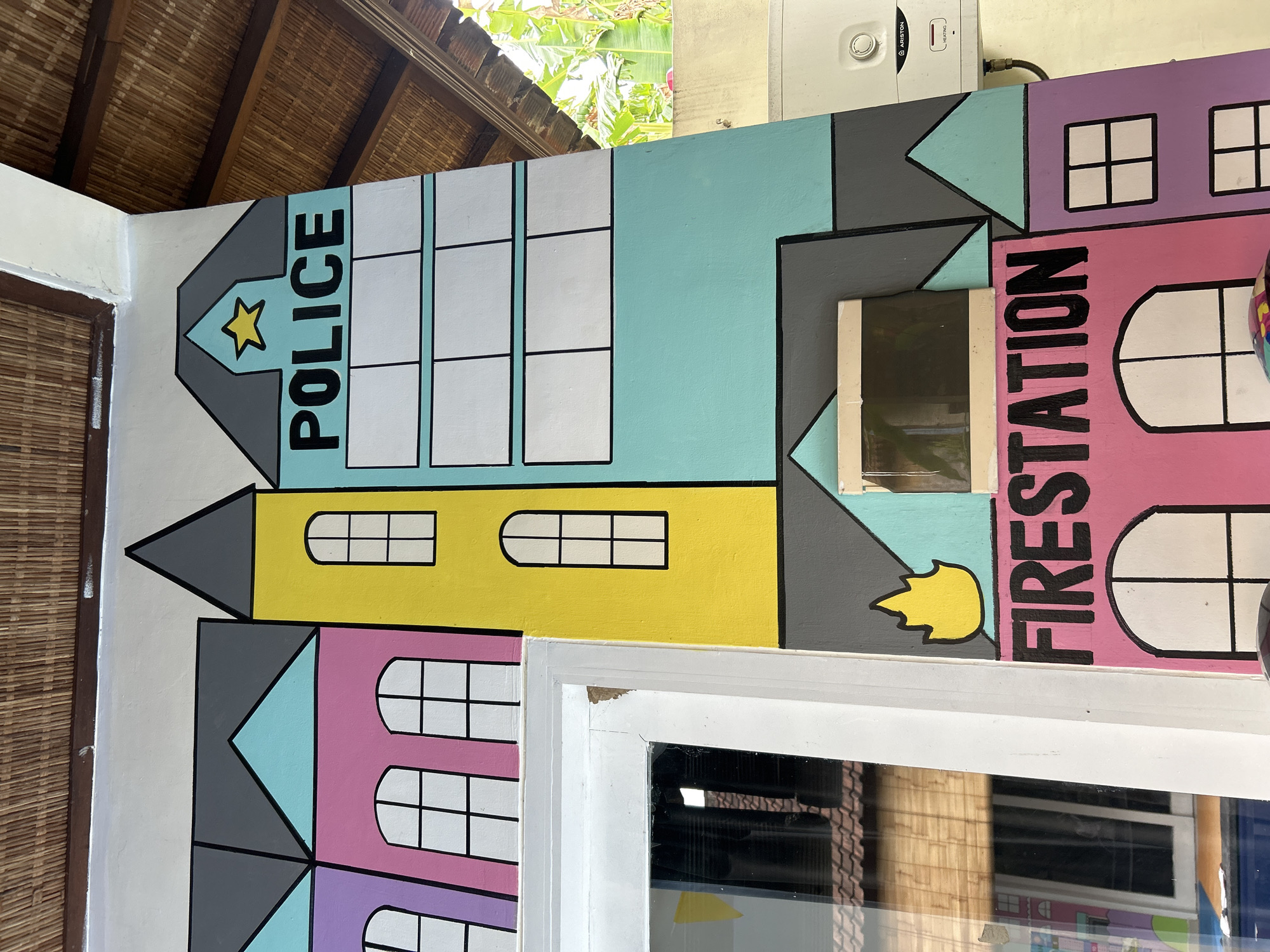 The image captures a colorful mural painted on an outdoor wall, likely in Kuta, Indonesia, given the context provided. The mural depicts stylized buildings, resembling a cityscape. On the right side, a section of a white Ariston water heater is visible, mounted on the wall above a turquoise building with a gray roof. The "POLICE" station is prominently featured with a yellow star above the text. Below this, a grid of empty white squares is painted. To the left and lower, a bright pink building with arched windows is labeled "FIRESTATION" in large black letters, with a yellow flame graphic above it. Other buildings in shades of purple, pink, and turquoise with black-lined windows fill the rest of the visible mural. A portion of a white-framed window is seen on the left side of the image, reflecting an interior space with what appears to be woven bamboo blinds. The ceiling above is covered with woven bamboo panels, suggesting an outdoor or semi-outdoor setting. The overall impression is a cheerful and whimsical depiction of community buildings.