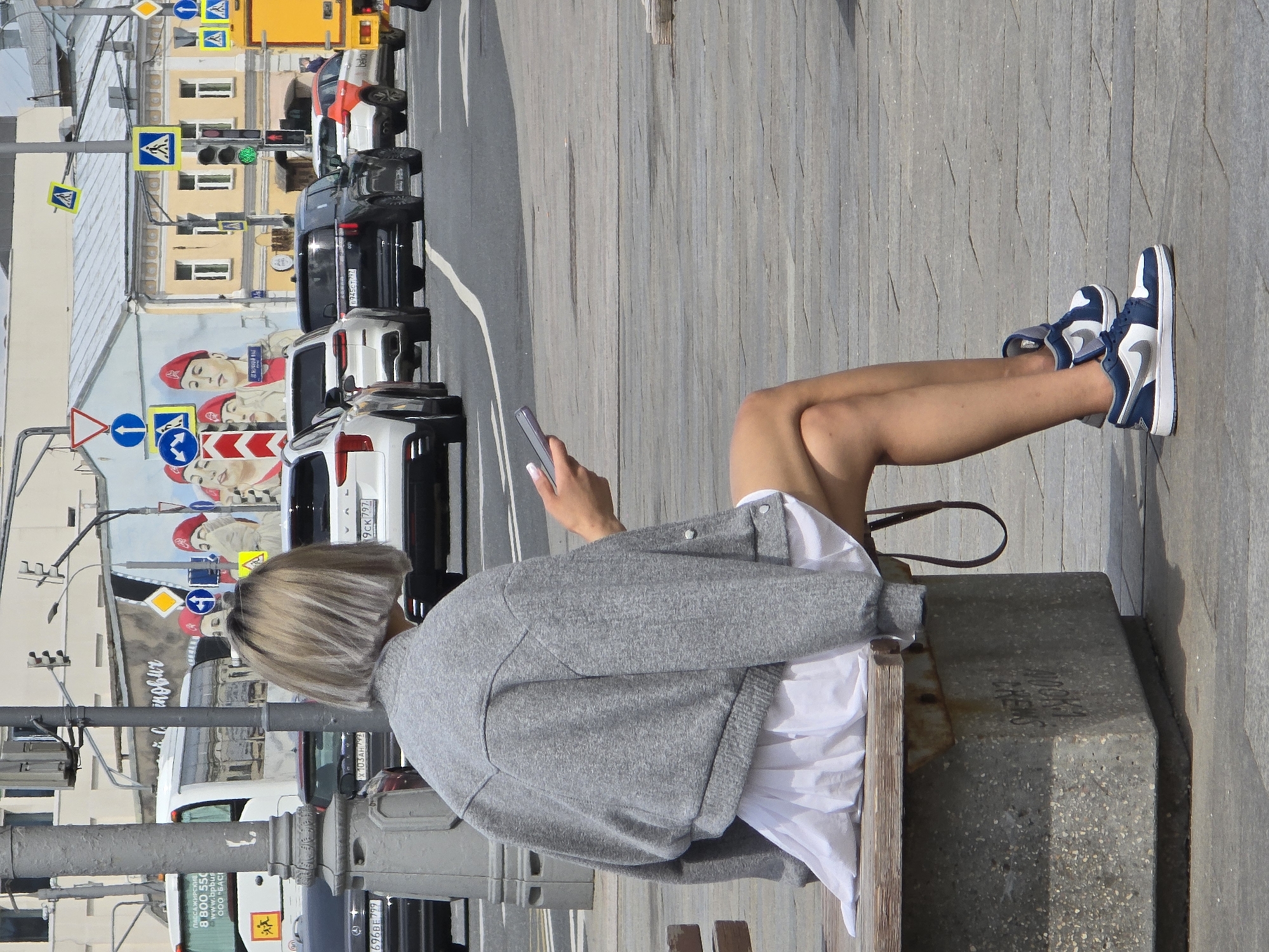 The image shows a woman sitting on a concrete bench in a street scene in Moscow, Russia. She is seen from the back, wearing a gray cardigan over a white shirt or dress, and blue and white sneakers. She is holding a smartphone. Behind her, there is a street with cars, street signs, and a building with a mural featuring images of people in red berets. The mural is visible behind her along with the street signs and traffic lights, hinting towards an urban setting, possibly in a busy street or square in Moscow. The cars are lined up as if at a stop, adding to the urban environment. The presence of a crosswalk sign and other directional signs point to a bustling city environment.